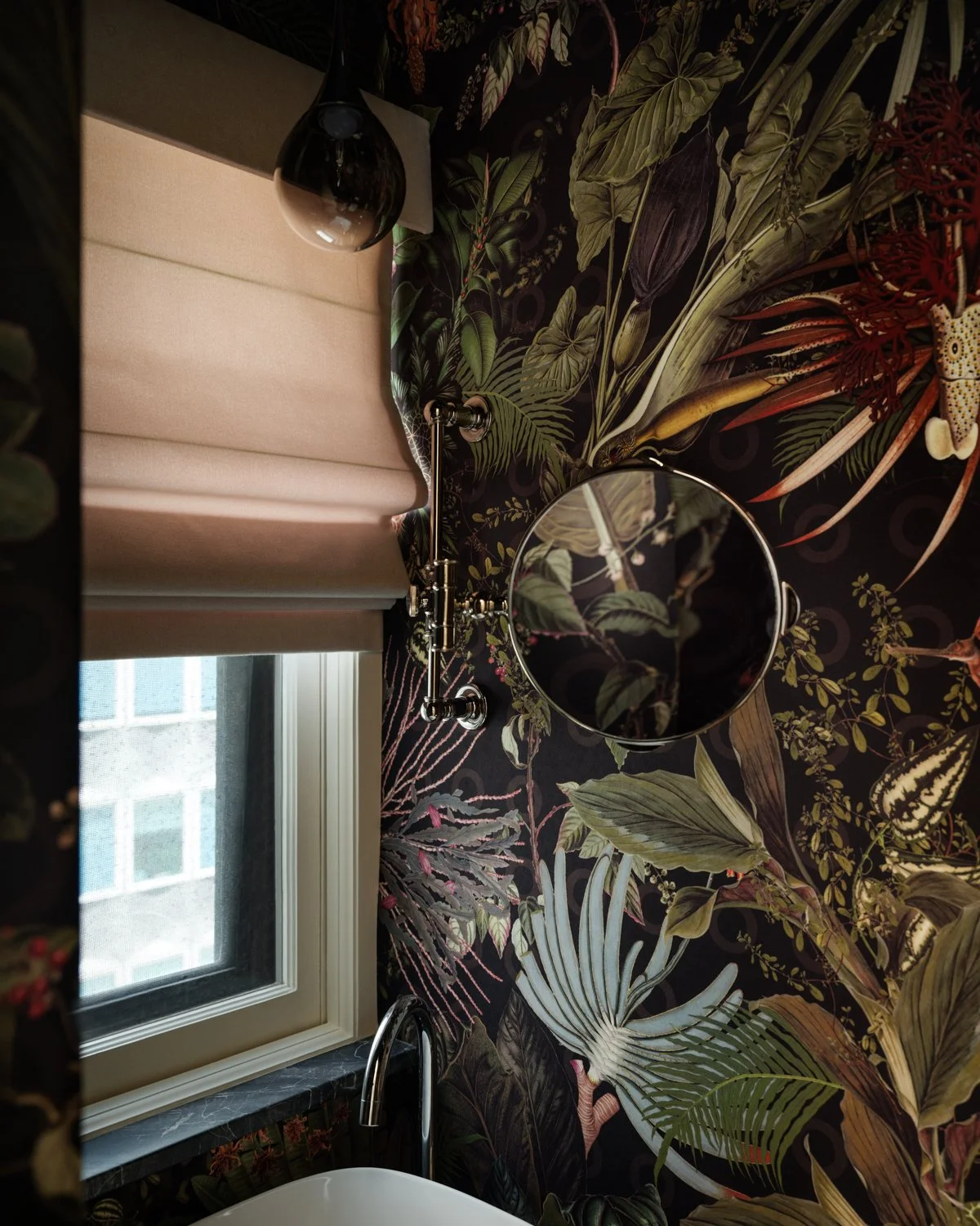 A bathroom with dark tropical leaf-patterned wallpaper, a window with a beige shade, a round mirror, a black hanging light, a white sink, and a faucet.