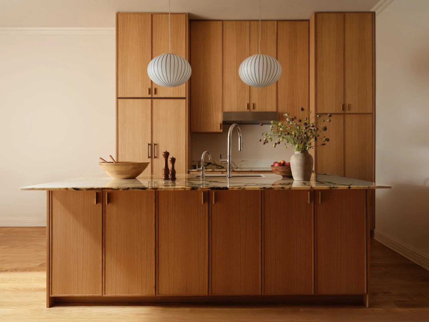 Modern kitchen with wooden cabinets, marble countertop, a vase of flowers, and two hanging pendant lights.