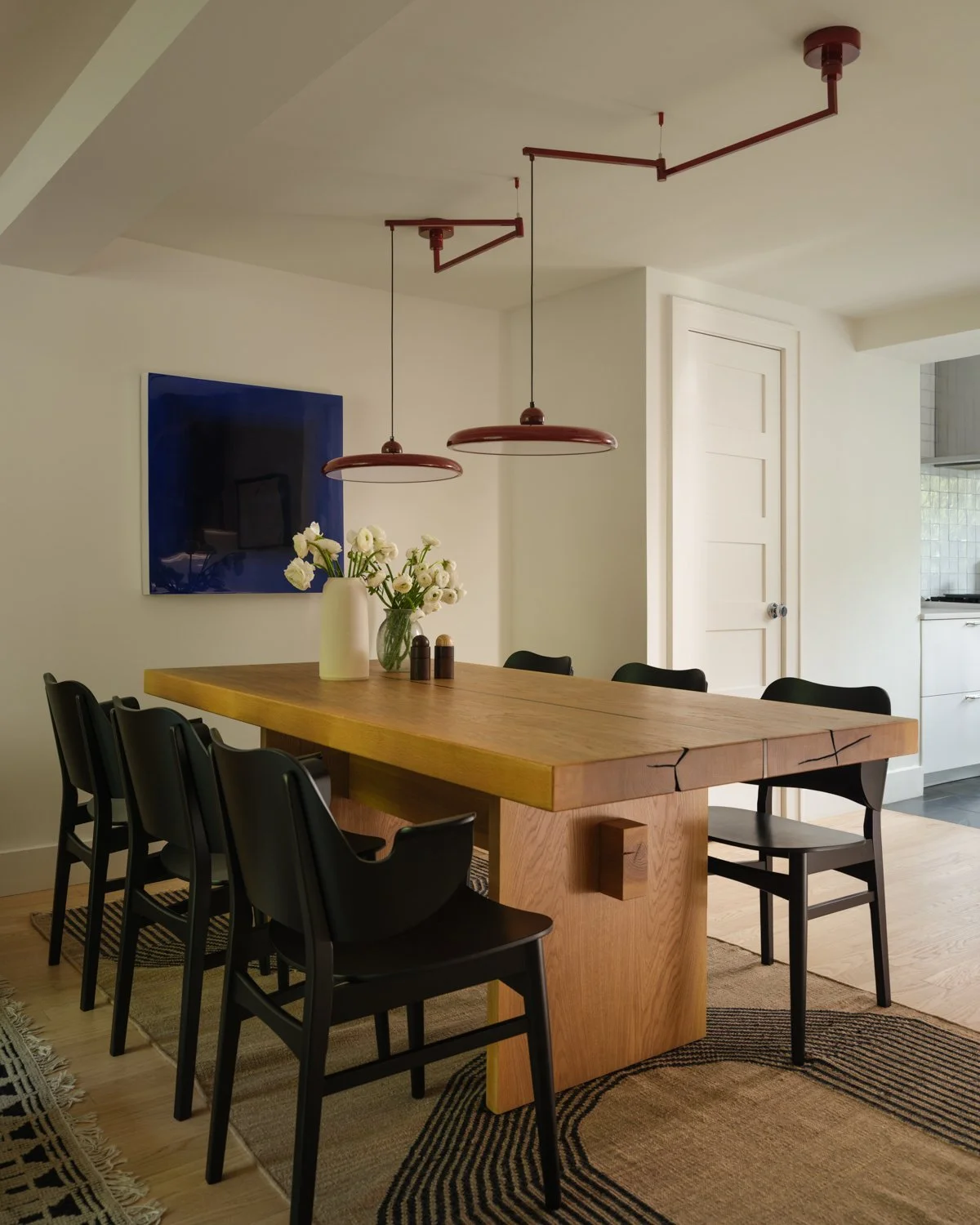 Dining room with a wooden table, six black chairs, a white wall with a mounted flat-screen TV, two red pendant lights hanging from the ceiling, a white door, and a vase with white flowers on the table.