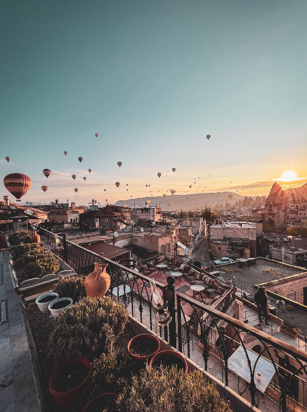 A rooftop view at sunset with numerous hot air balloons floating in the sky over a town area with traditional buildings.