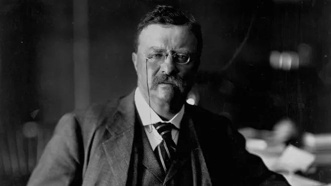 Black and white photograph of a man with glasses, a mustache, and beard, wearing a suit and tie, seated indoors.