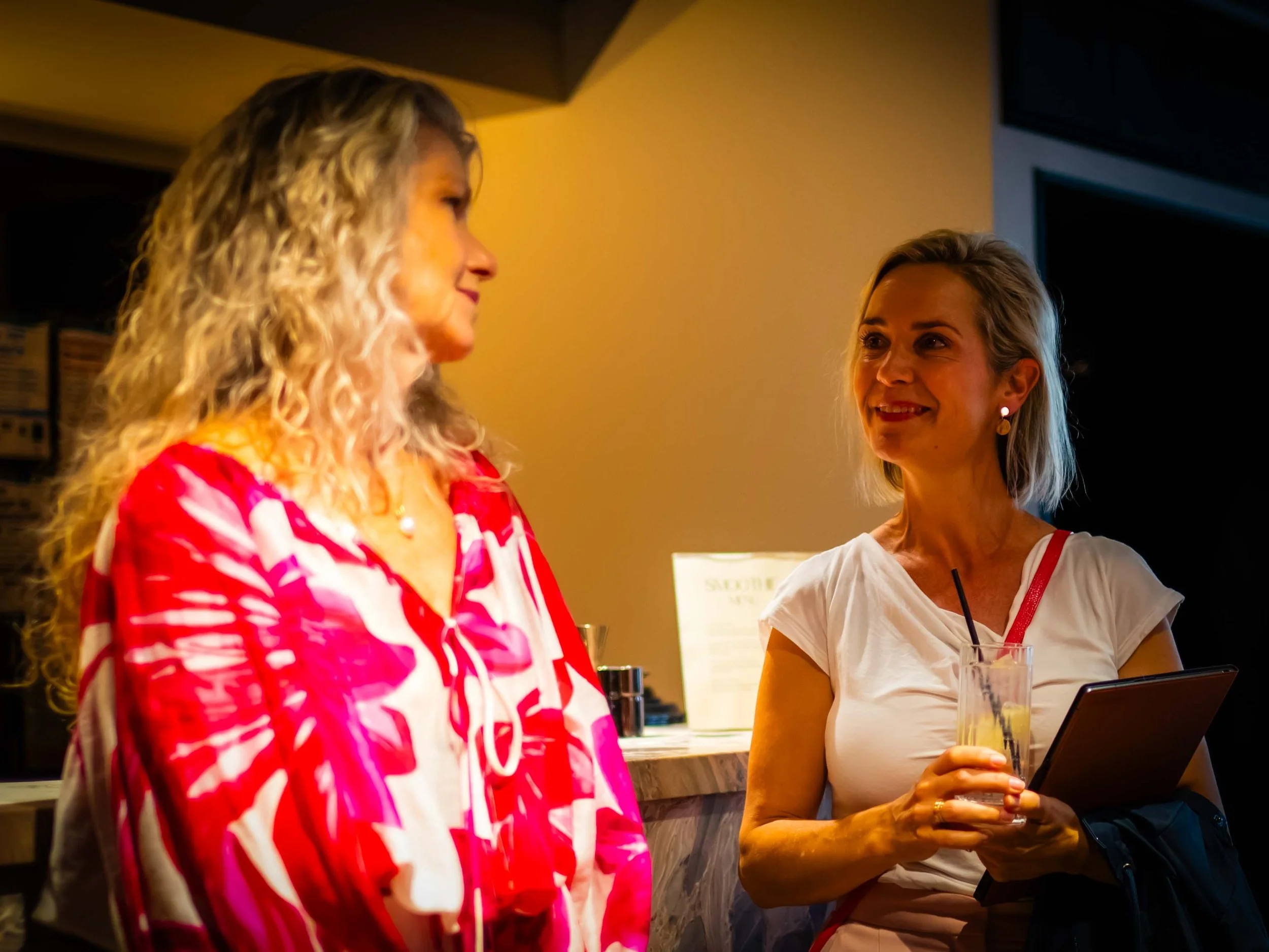 Two women having a conversation indoors. One woman is in a red and white patterned top with blond curly hair; the other is in a white t-shirt with blond straight hair, holding a tablet and a drink.