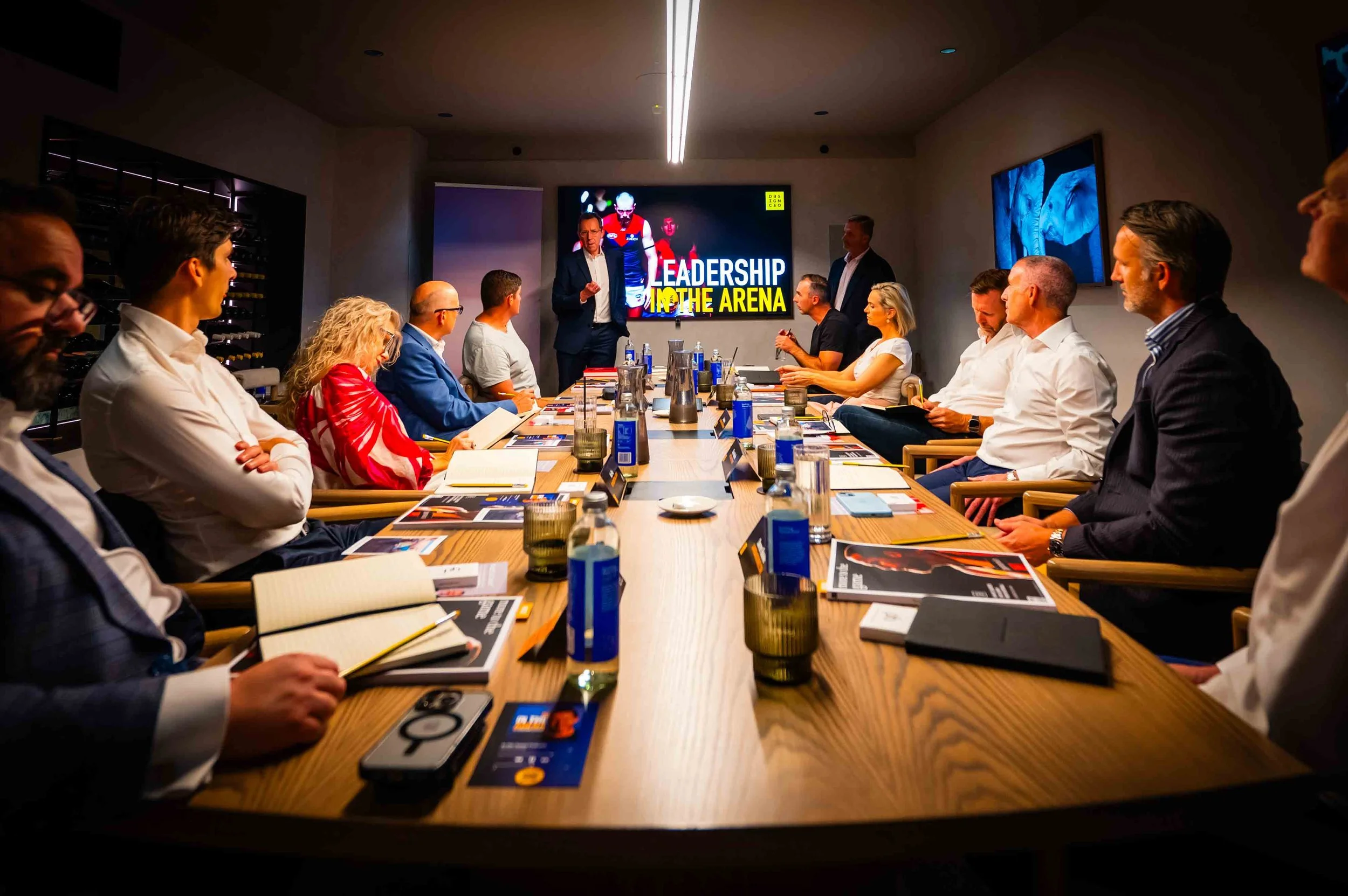 Business meeting room with approximately twelve diverse professionals seated around a long conference table, a man standing and speaking at the head of the table, a large screen behind him displays 'Leadership in the Arena,' and the room is equipped with notebooks, water bottles, and electronic devices.