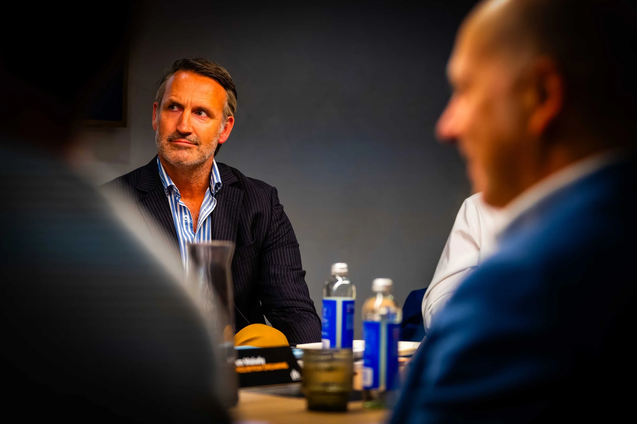 Business meeting with a man in a dark pinstripe suit and blue striped shirt in focus, seated at a table with water bottles and glasses, listening intently.