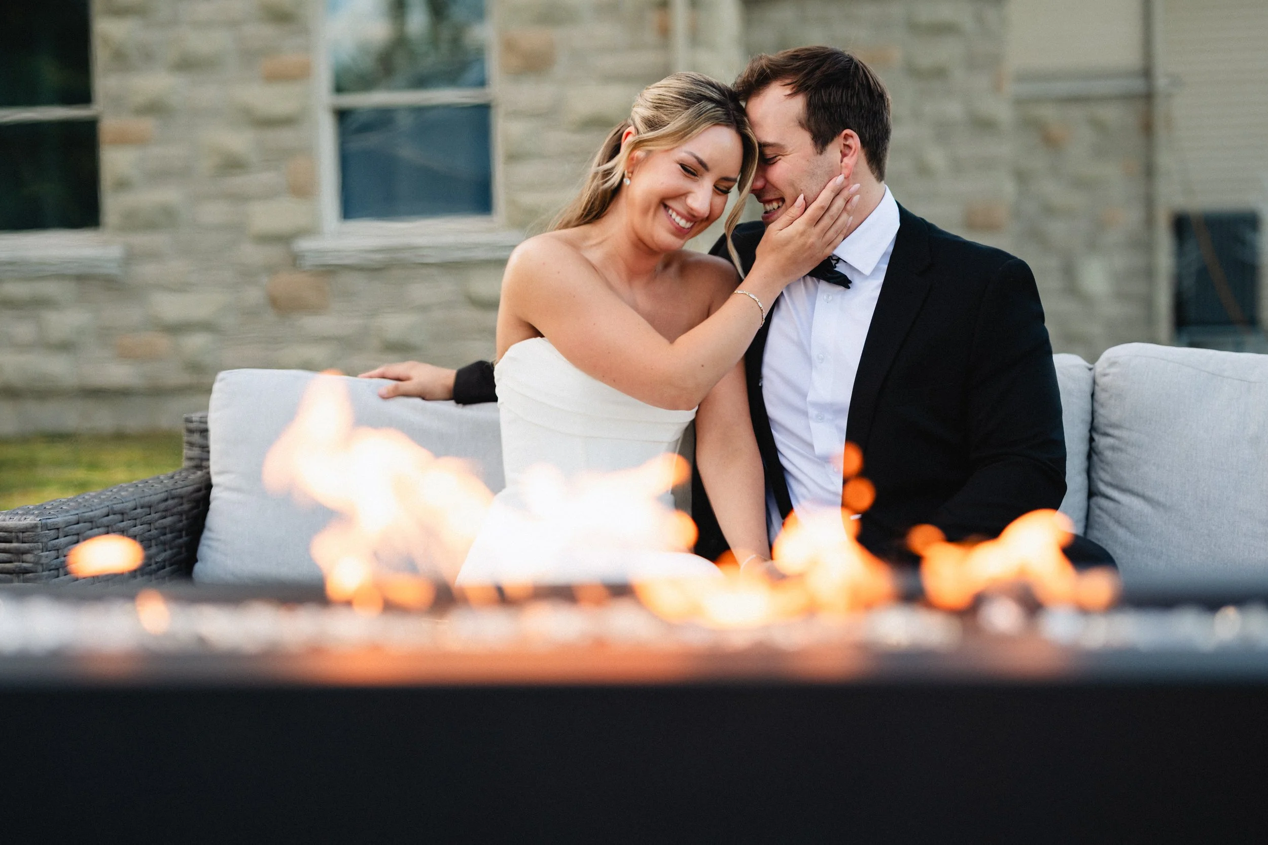 A bride and groom sitting close together on an outdoor sofa, smiling and touching foreheads, with a fire pit in the foreground and a house with stone walls in the background.