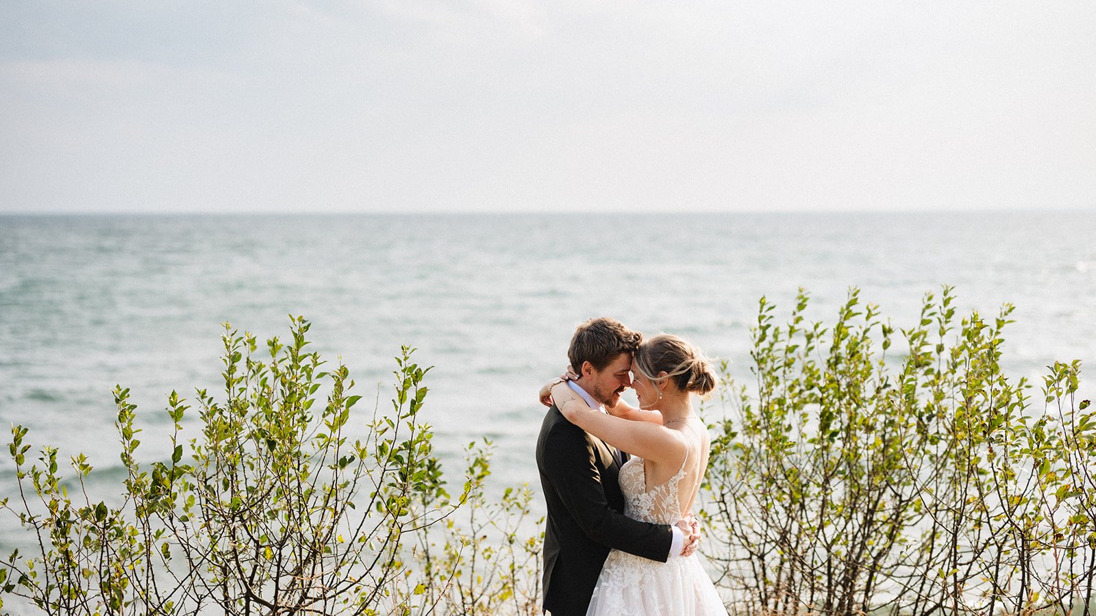 A couple in wedding attire hugging on a beach with bushes and the ocean in the background.