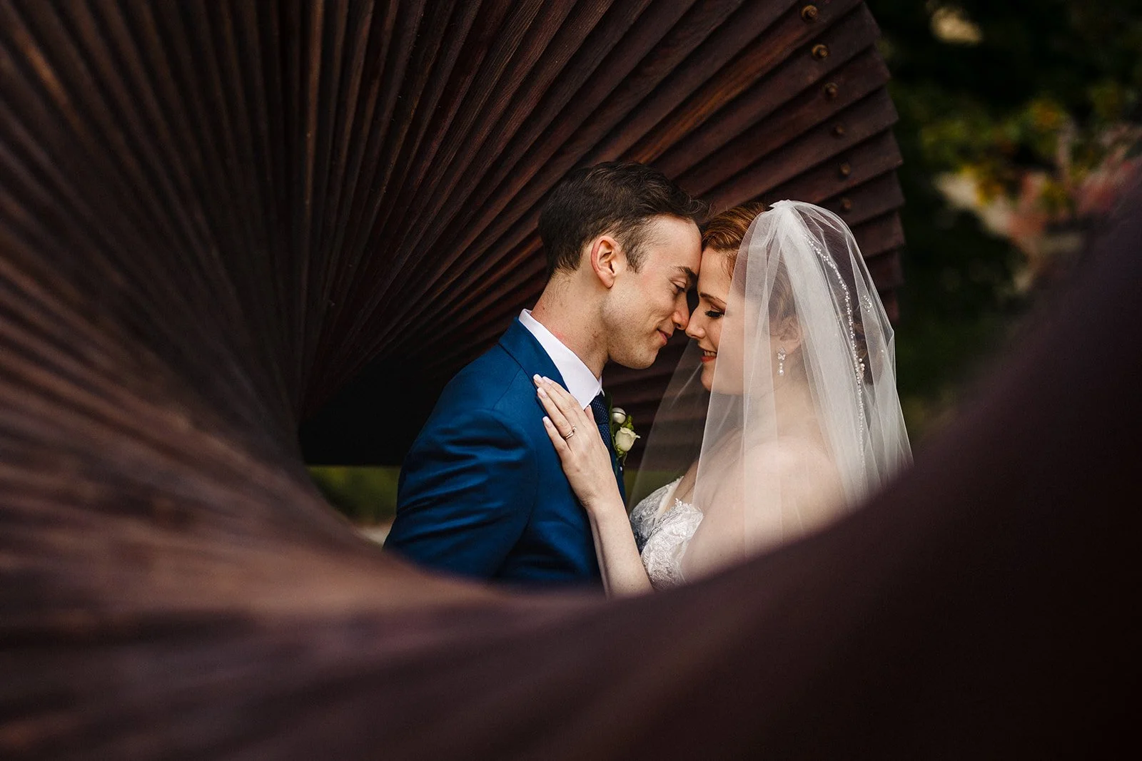 A bride and groom close together with their foreheads touching, smiling at each other, framed by a large, dark brown umbrella.