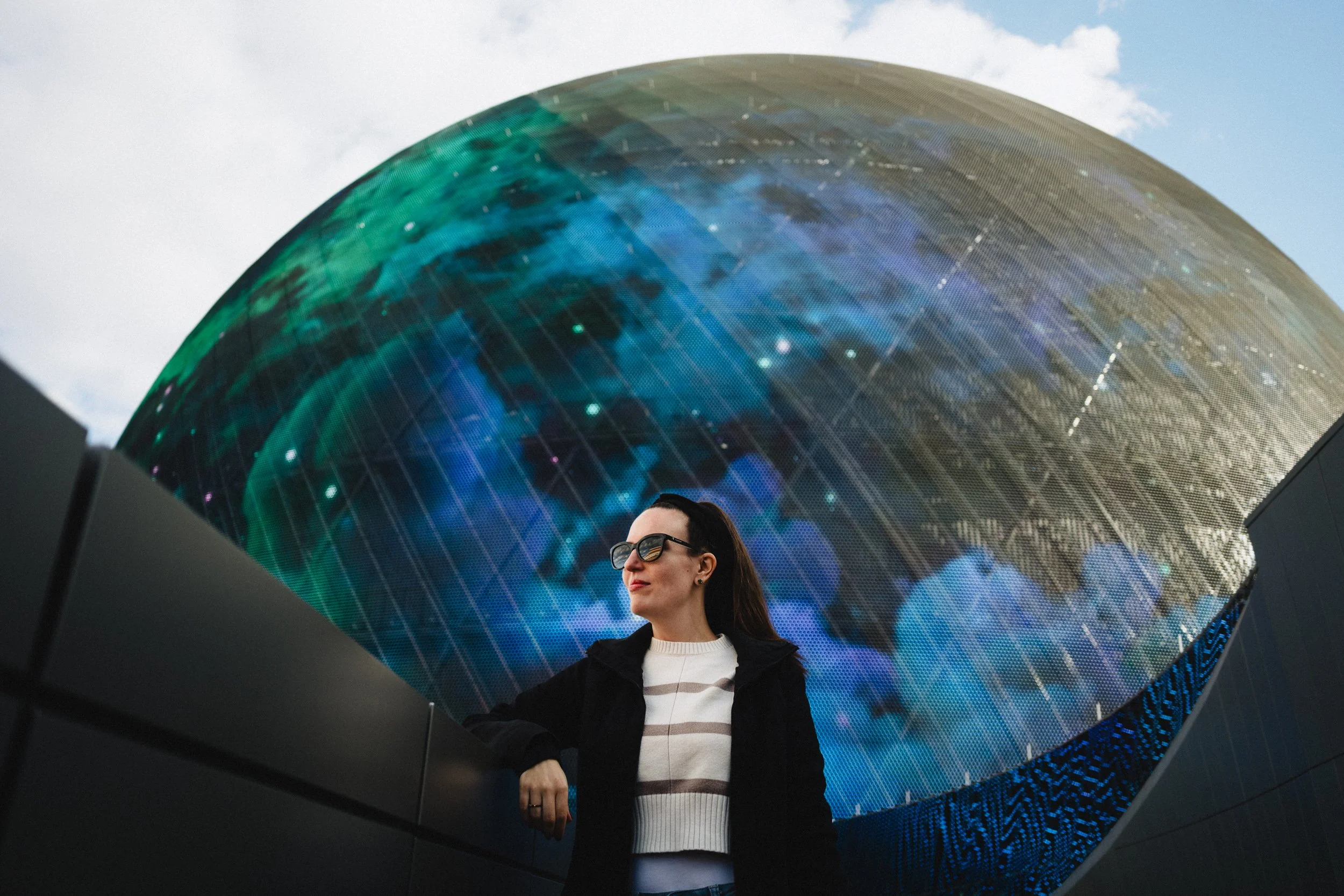 A woman wearing sunglasses and a black jacket standing in front of a large, curved digital display showing a colorful, abstract image of Earth from space.