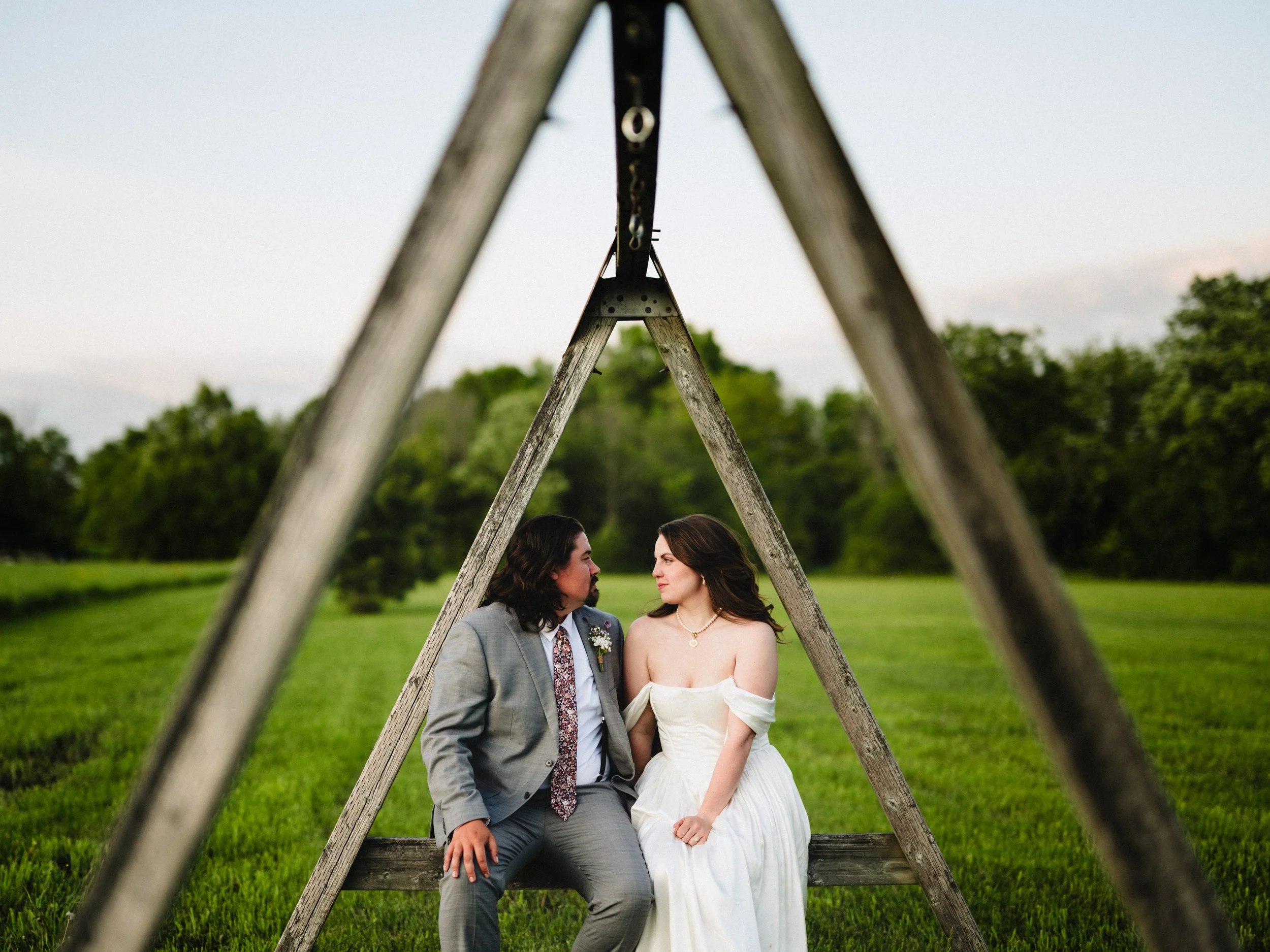 A couple sitting on a wooden bench under a large wooden A-frame structure in a grassy field, with trees in the background, during sunset.