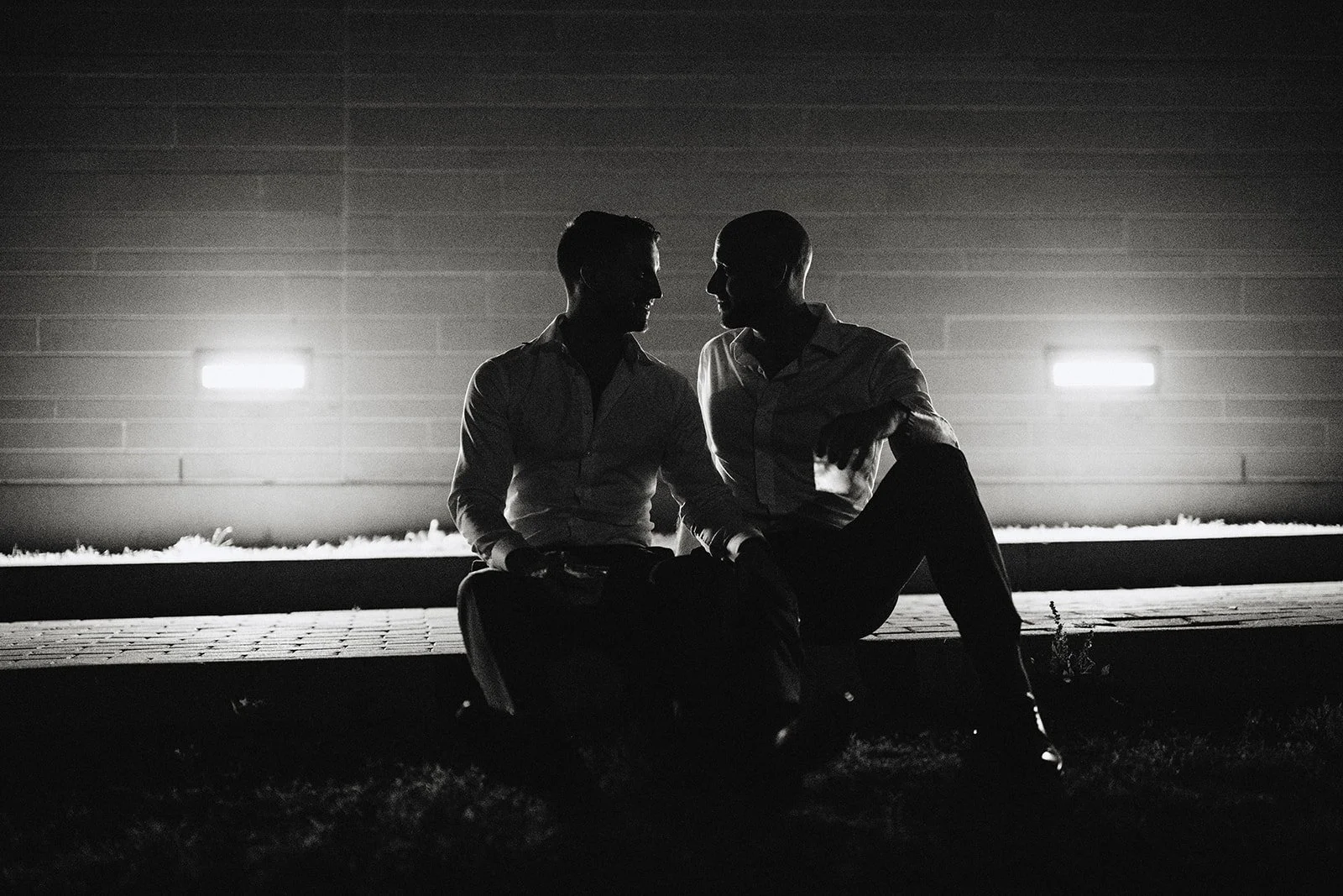 Two men sitting close together on a curb at night, silhouetted against bright lights in the background.