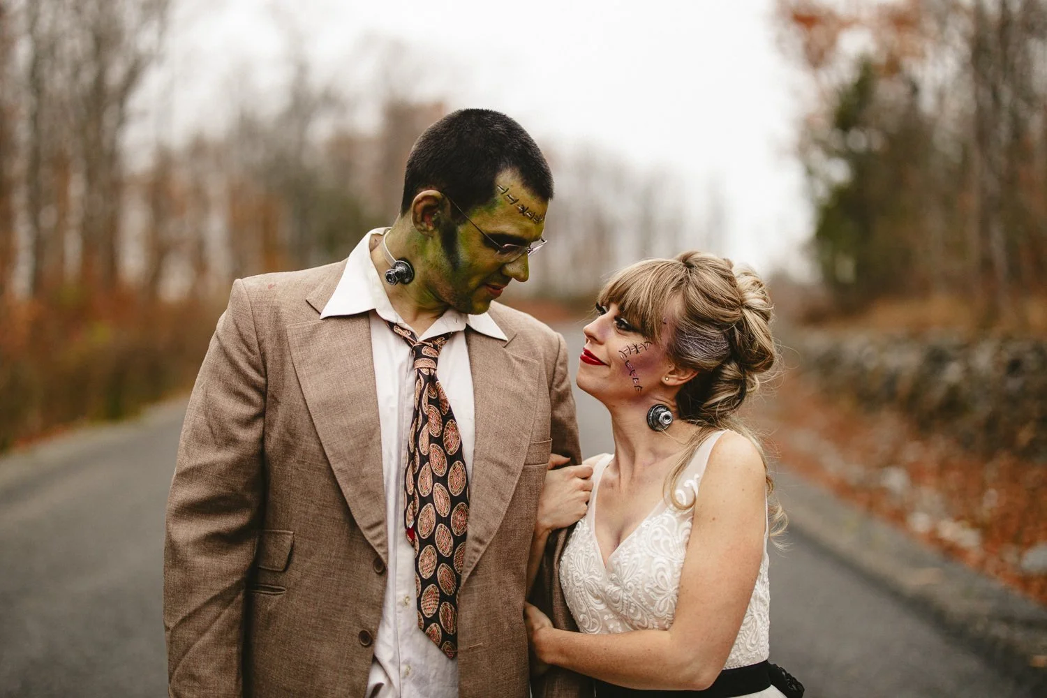 A couple in costume with fake scars and face paint, standing outdoors on a road with autumn trees in the background. The man is wearing a suit and tie, and the woman is in a white dress with black accents, both with relaxed expressions and close to each other.