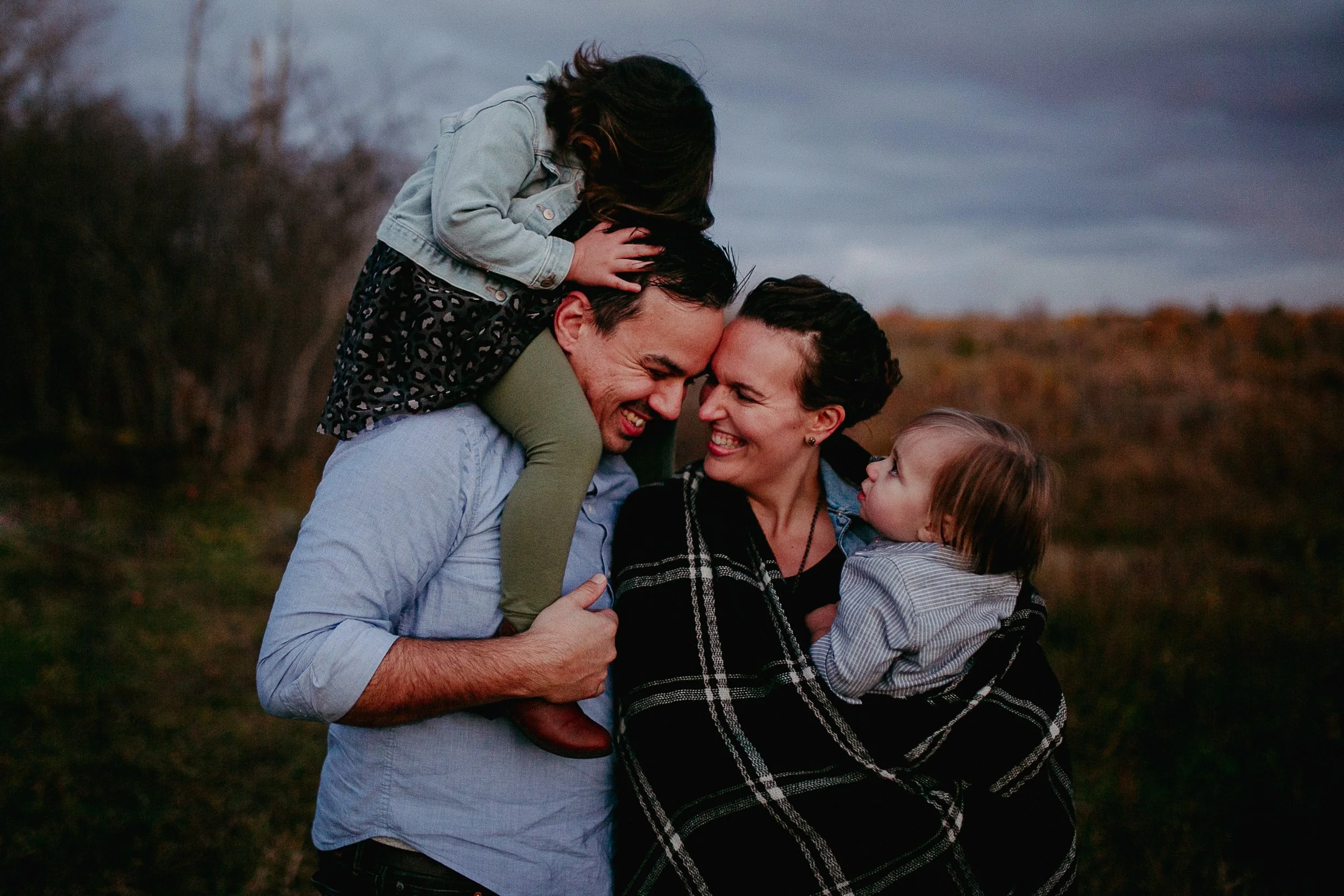 A family outdoor portrait with two parents and two children, embracing and smiling warmly at each other during sunset or twilight.