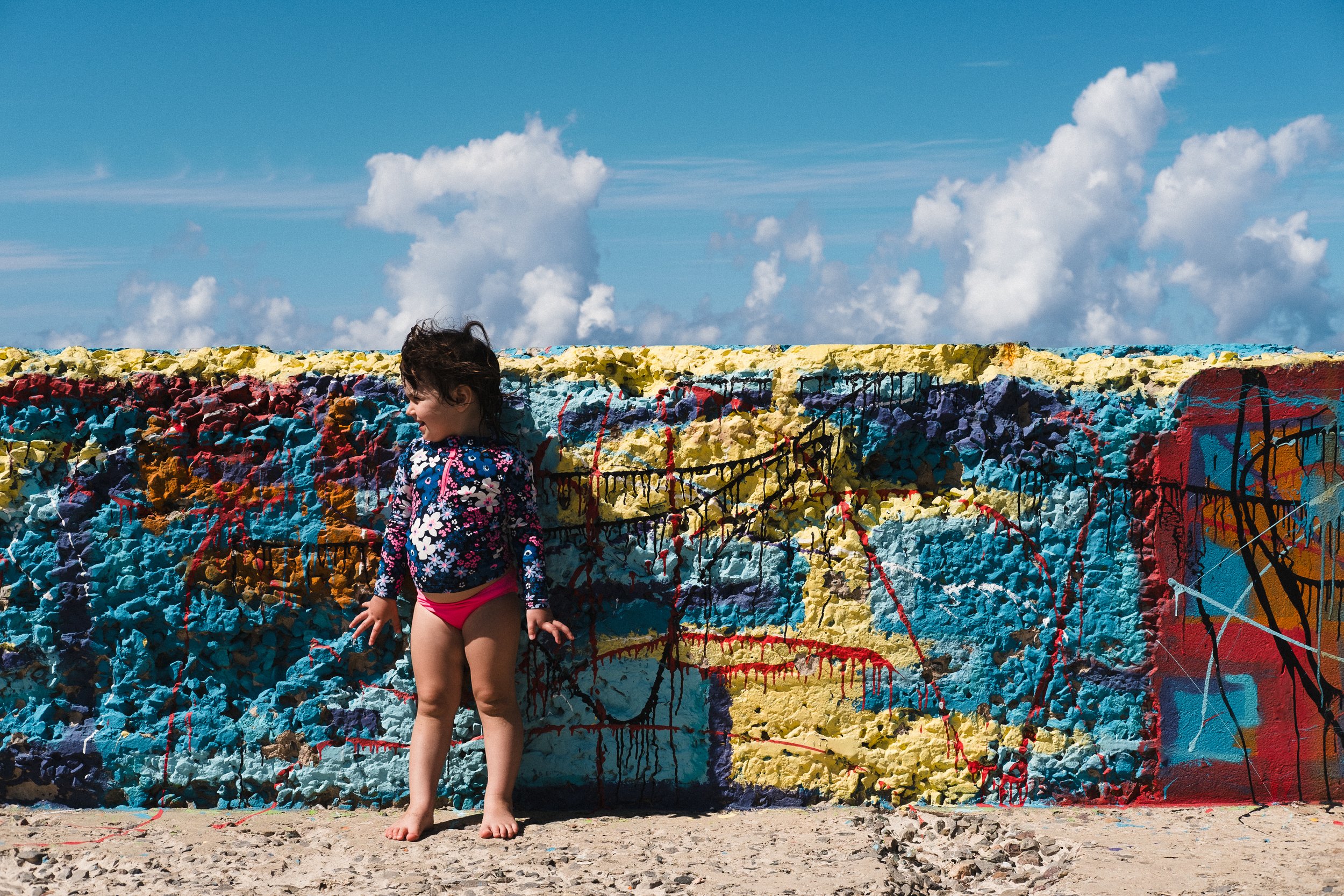 Young girl in a floral jacket and pink shorts standing barefoot in front of a colorful, graffiti-covered concrete wall under a blue sky with clouds.