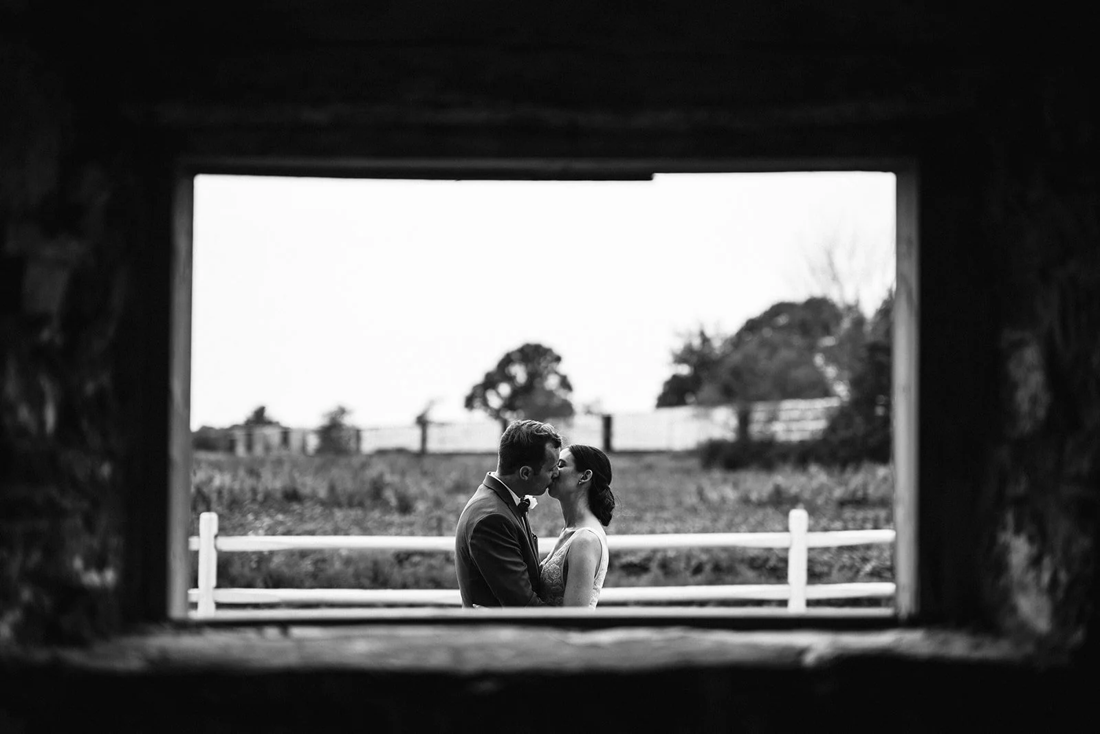 Black and white photo of a couple kissing outside, viewed through a window or opening with a wooden frame, with trees and a fence in the background.