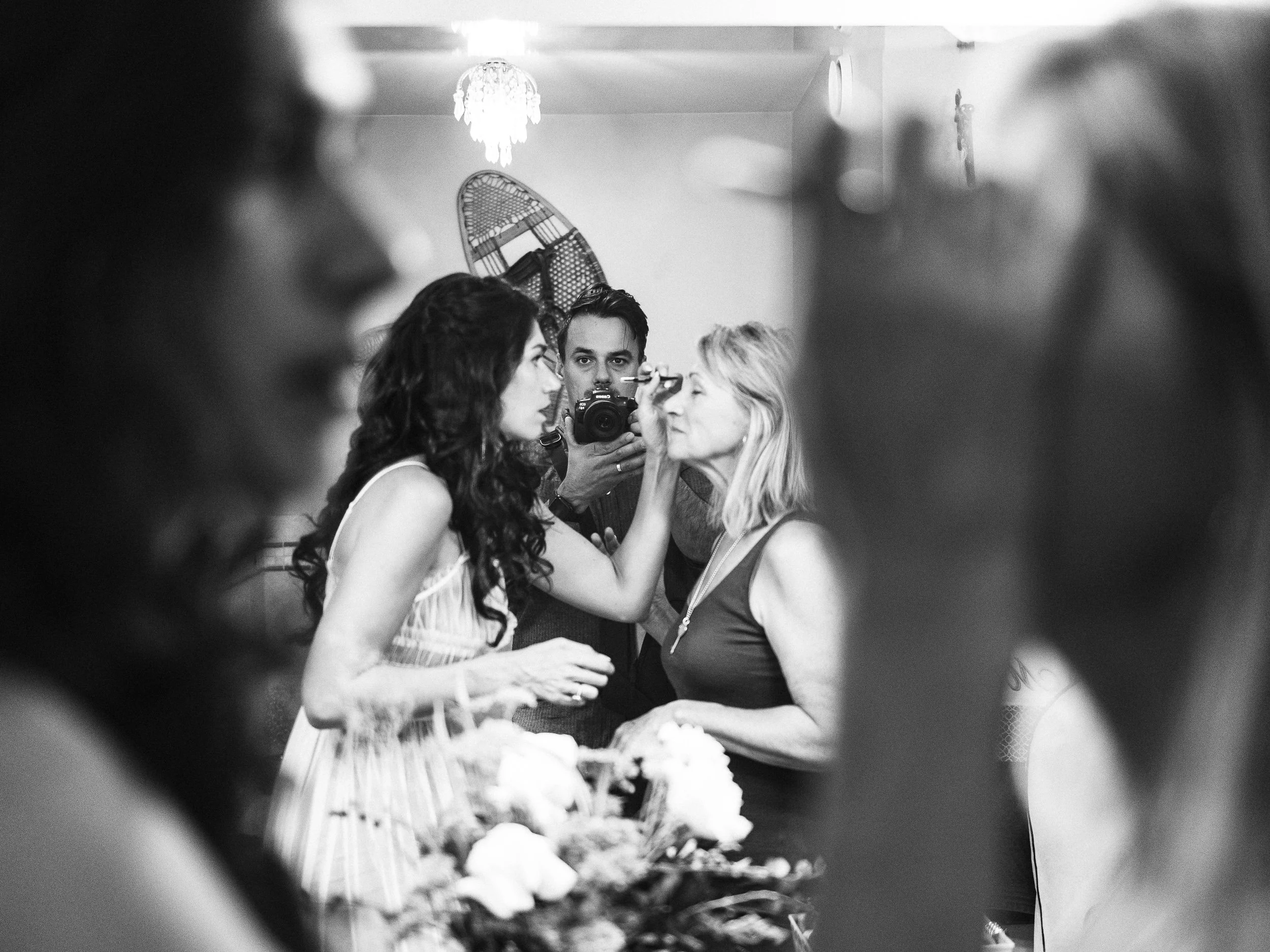 A woman with dark, curly hair receives makeup from a woman with blonde hair, at an indoor gathering, with a photographer capturing the moment in the background.