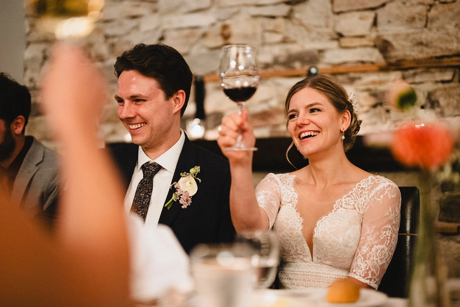 A bride and groom sitting at a wedding reception, smiling as the bride raises a glass of red wine.