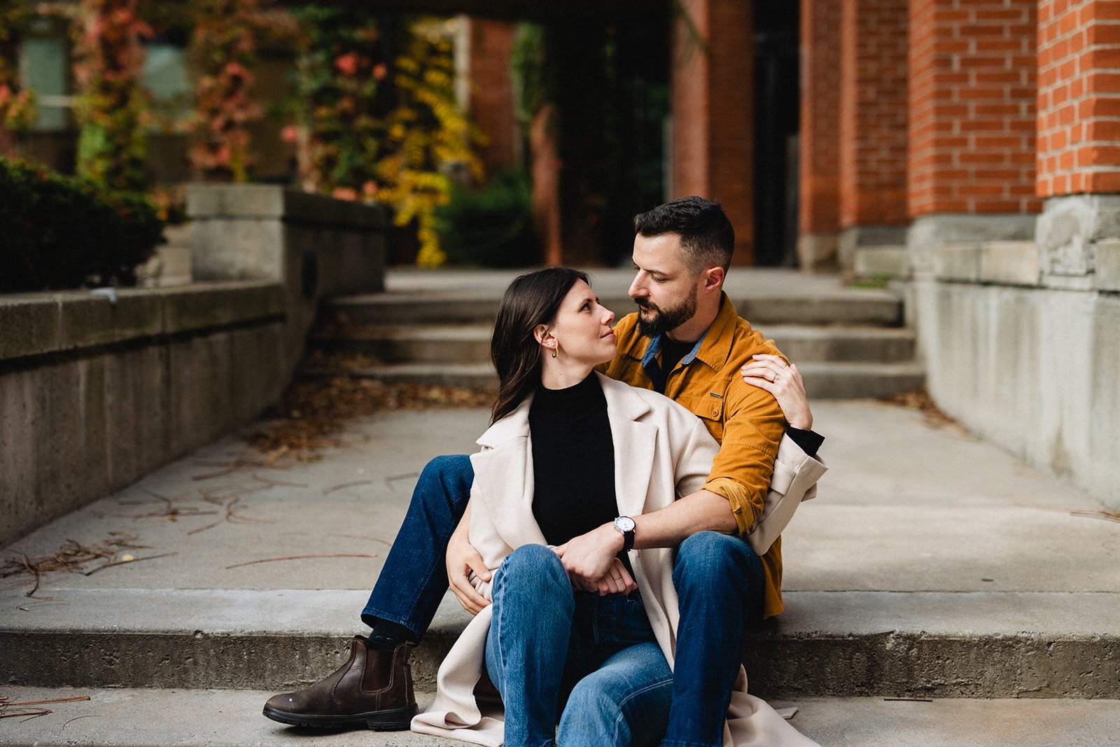 A couple sitting on steps outdoors, looking into each other's eyes, with trees and brick building in the background.