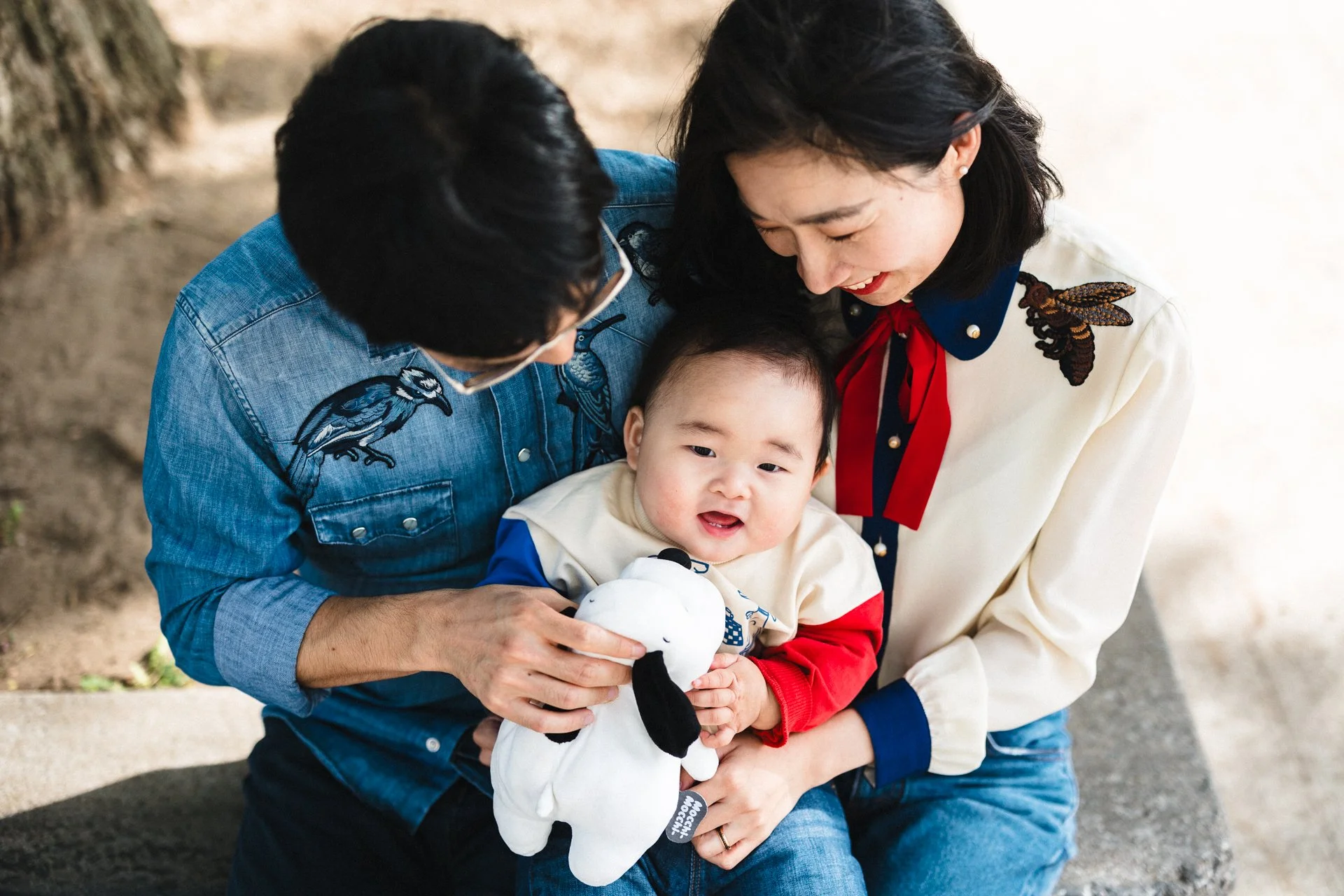 A family of three, including a man, a woman, and a baby, sitting outdoors on a concrete surface. The baby is holding a stuffed cow toy, and everyone is smiling and engaging with each other.