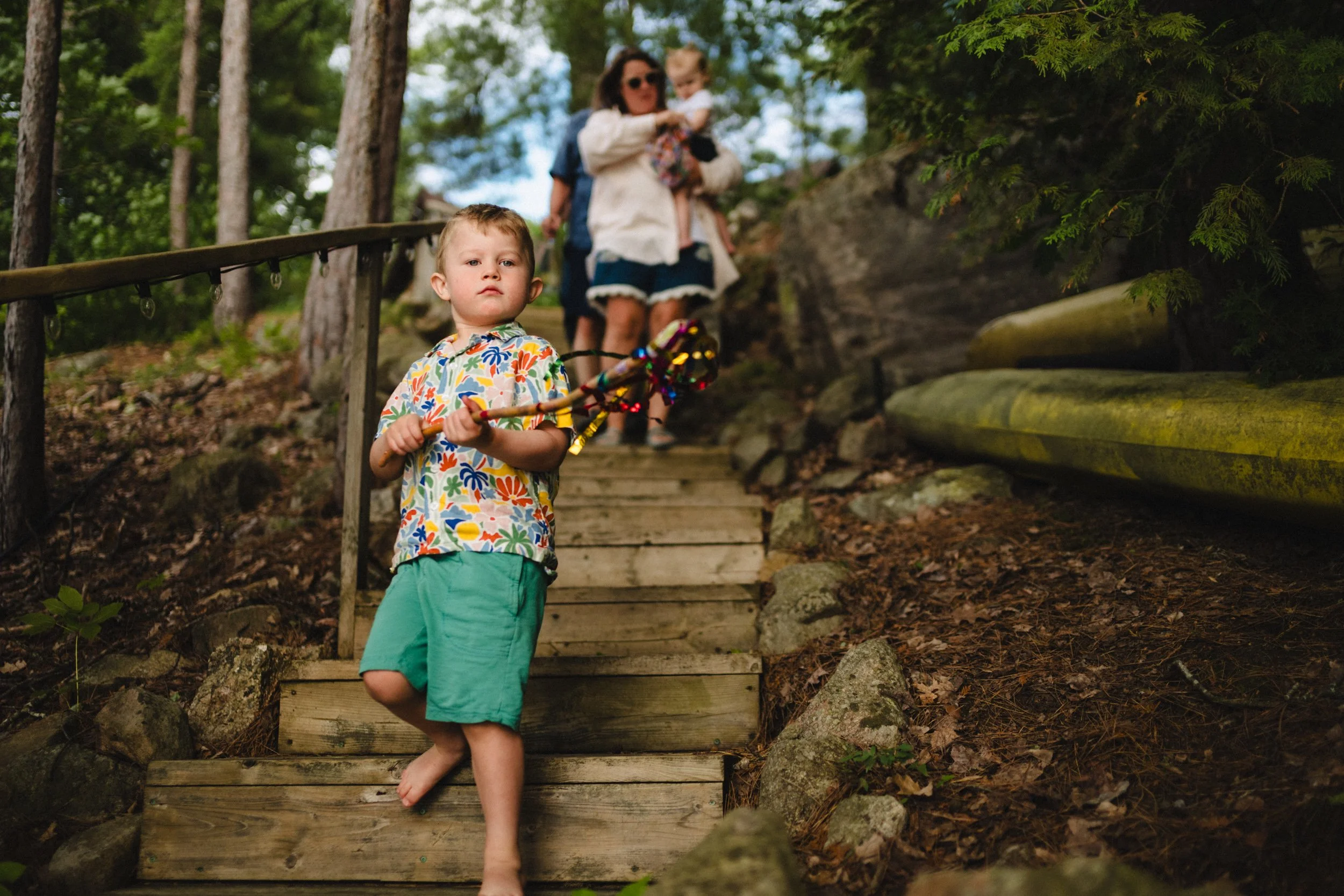 A young boy with a colorful shirt and green shorts holding a stick, walking down wooden stairs outdoors, with a woman and another child in the background on a wooded hillside.