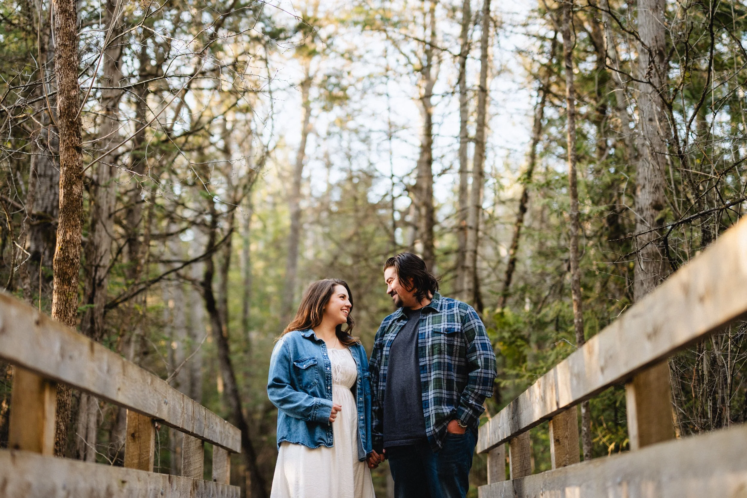 A couple standing on a wooden bridge in a forest with tall trees, holding hands, smiling, and looking at each other.