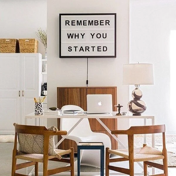 Home office space with a white desk, two wooden chairs with cushions, a laptop, a table lamp, and a sign on the wall reading "REMEMBER WHY YOU STARTED."