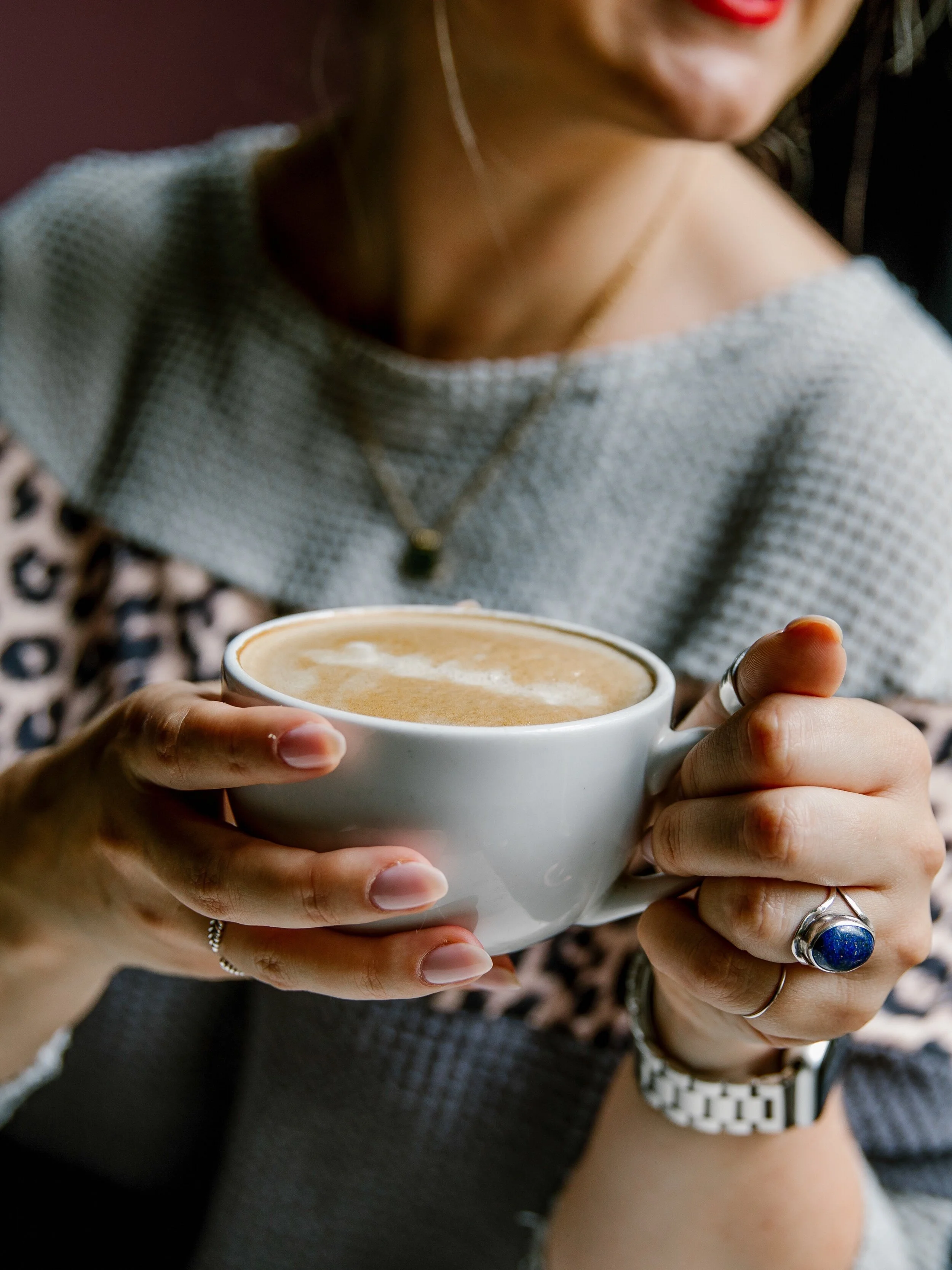 D holding a white coffee mug with latte art, wearing a silver ring with a blue stone, a silver ring, and layered bracelets. Part of their face with red lipstick is visible, and they are dressed in a gray sweater with an animal print shirt underneath.