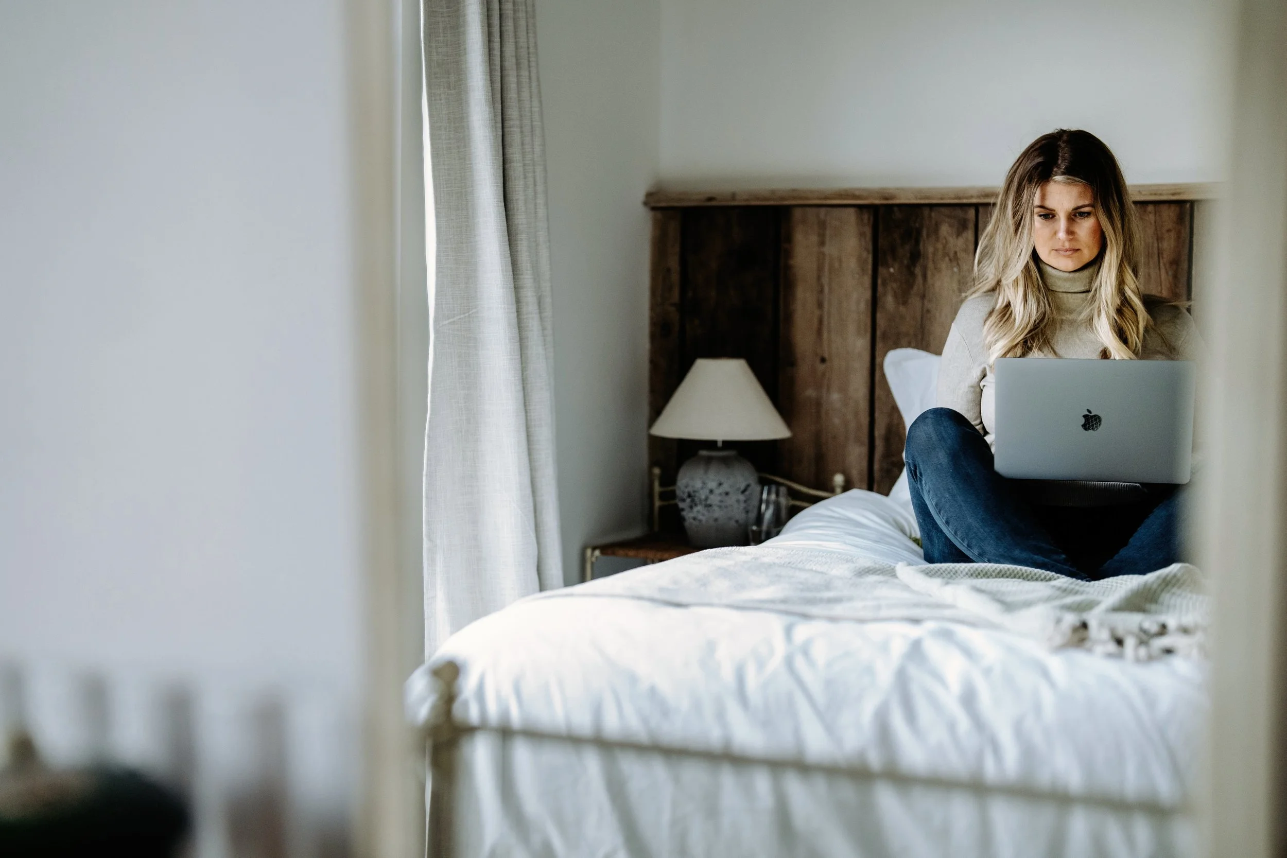 Dalia Courridge - A woman with blonde hair sitting on a bed with a laptop, in a bedroom with neutral walls, a wooden headboard, a lamp, and curtains.