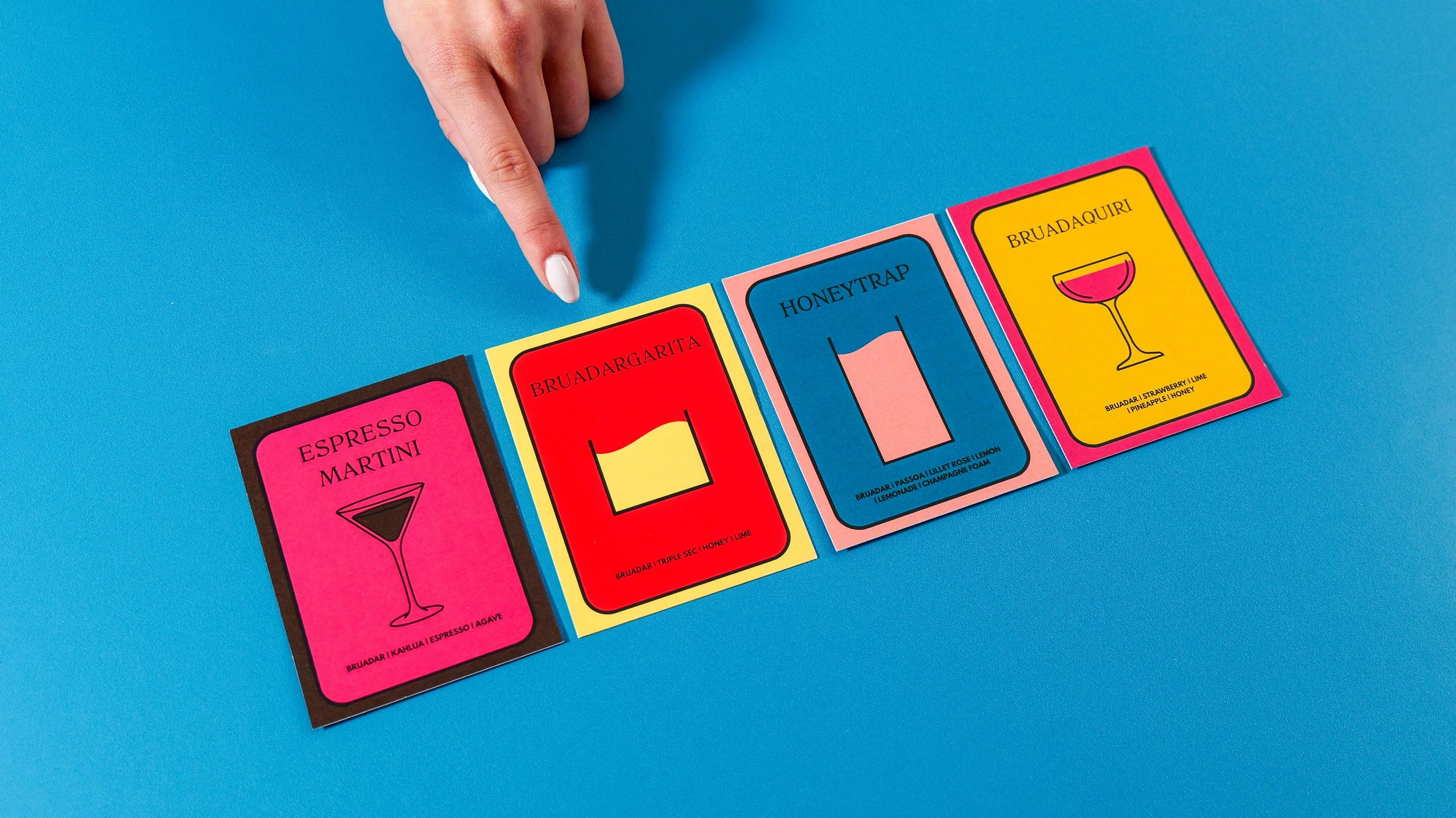 Colorful cocktail recipe cards laid out on a blue surface, with a finger pointing at the cards.