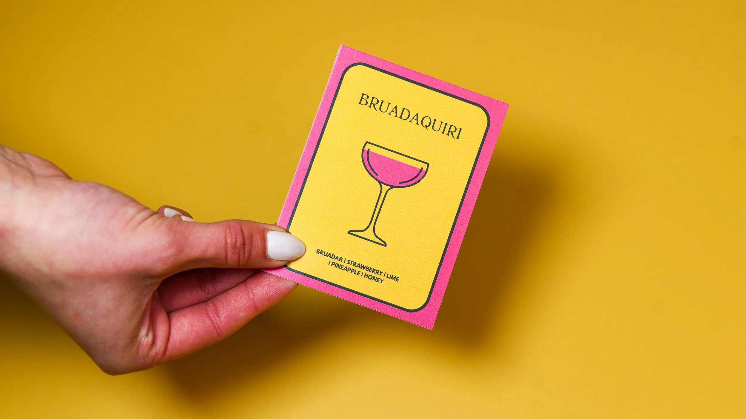 A hand holding a colorful card with a pink border and yellow background, featuring a line drawing of a cocktail glass and the words "BRUADAQUIRI" along with ingredients listed below.