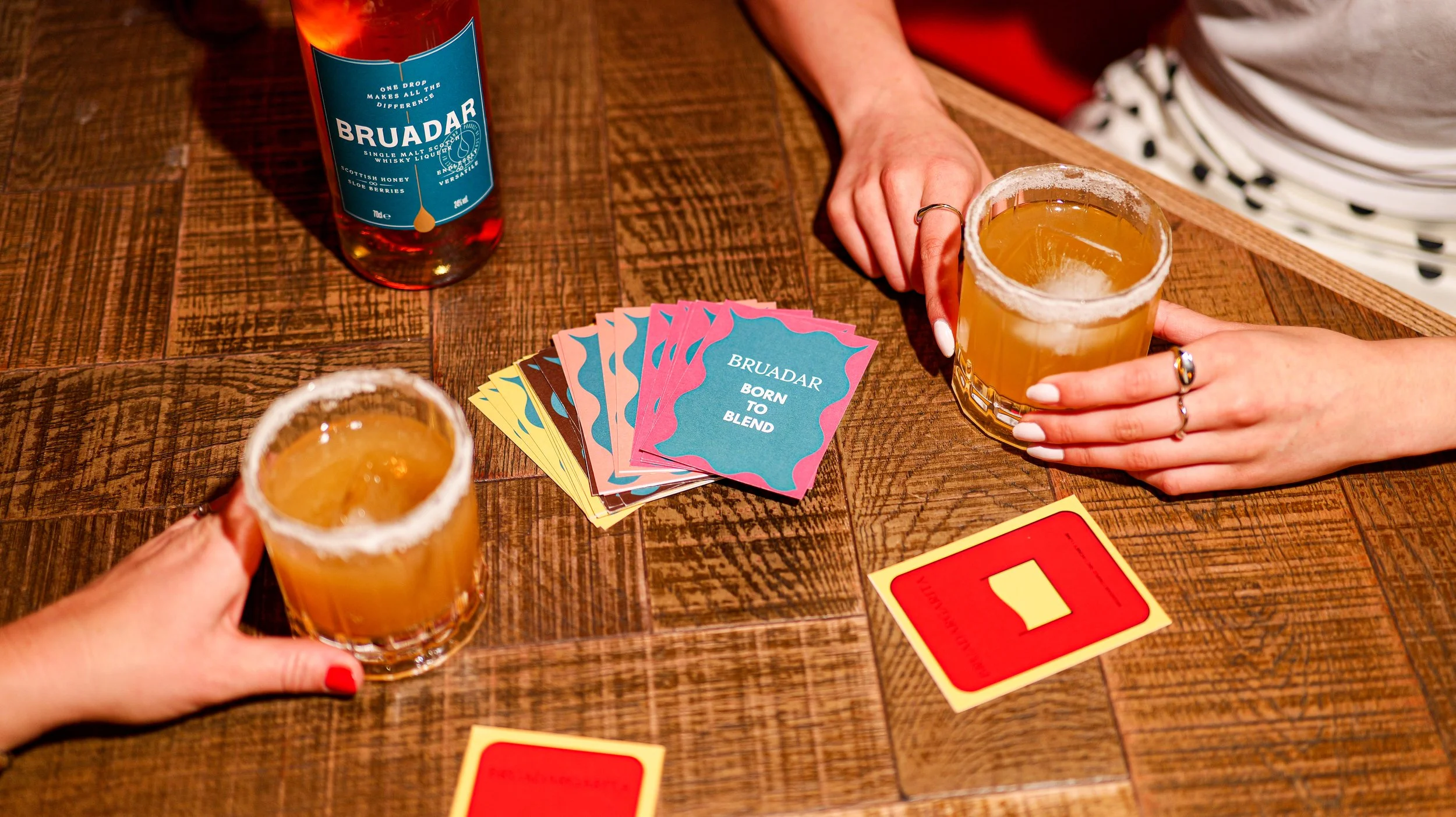 Two women holding drinks with salted rims, playing a card game on a wooden table, with a bottle of Bruadar whisky and colorful cards that read 'Born to Blend' in the background.