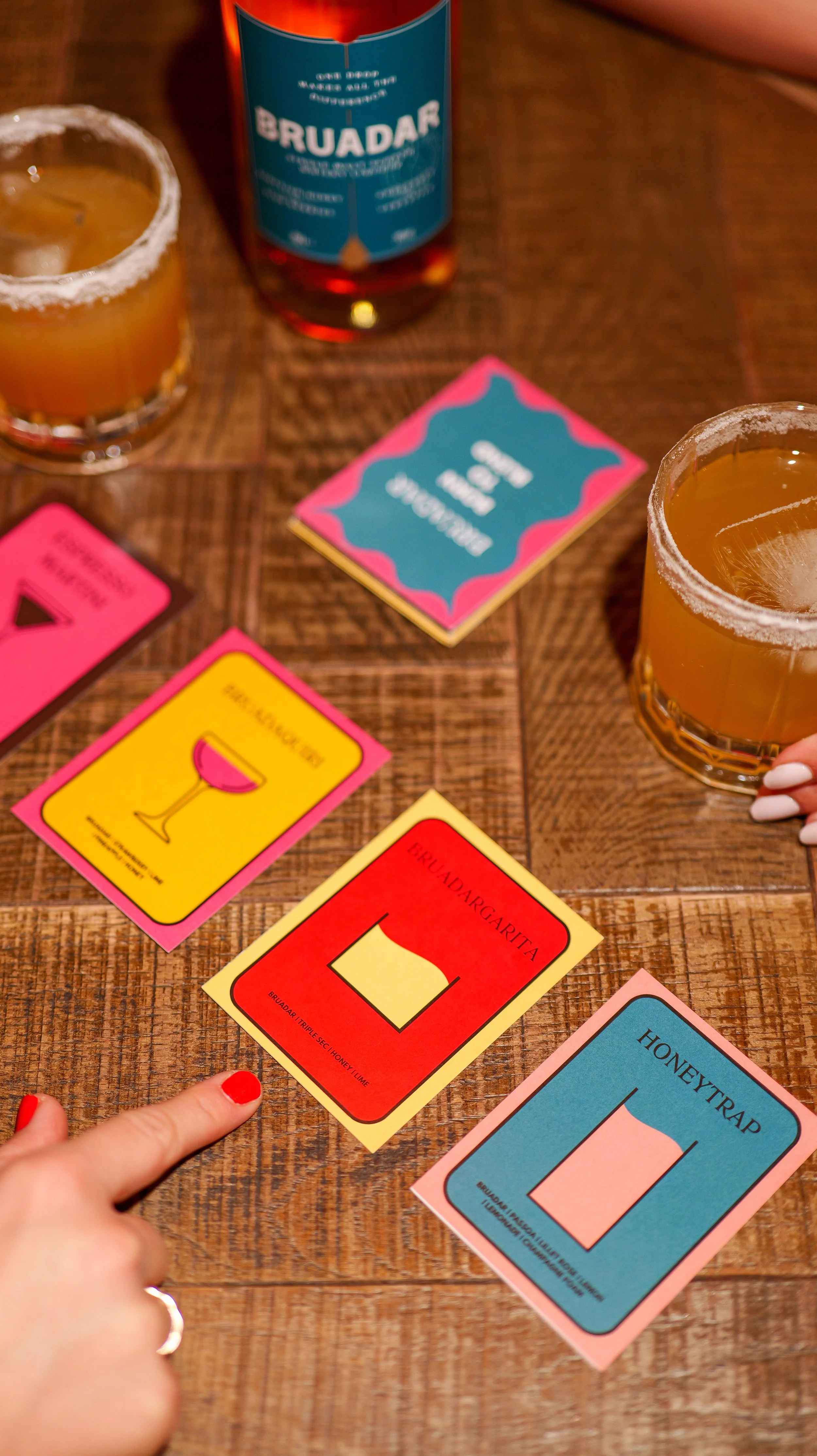 Colorful cocktail recipe cards on a wooden table with drinks and a bottle of alcohol in the background.