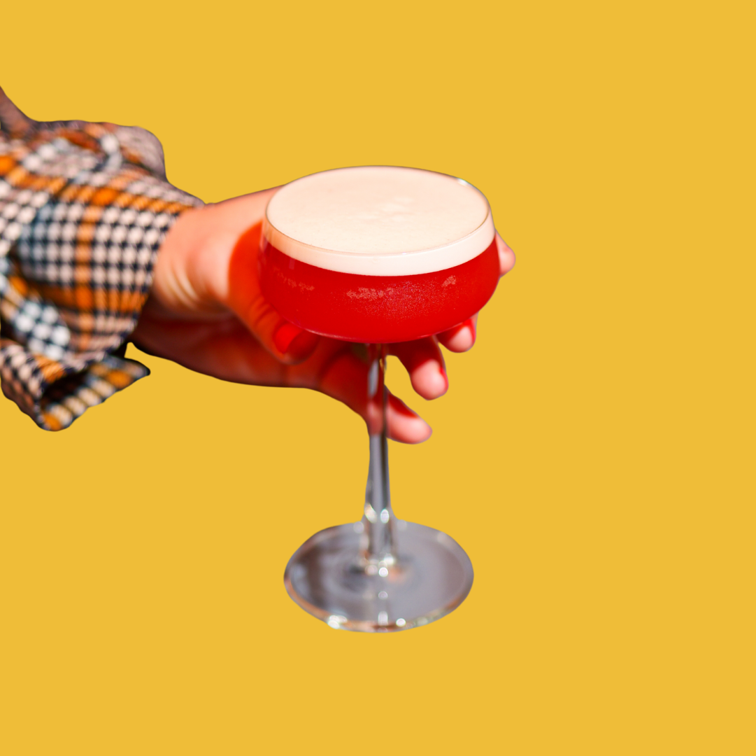Person holding a red cocktail glass with frothy drink against yellow background.