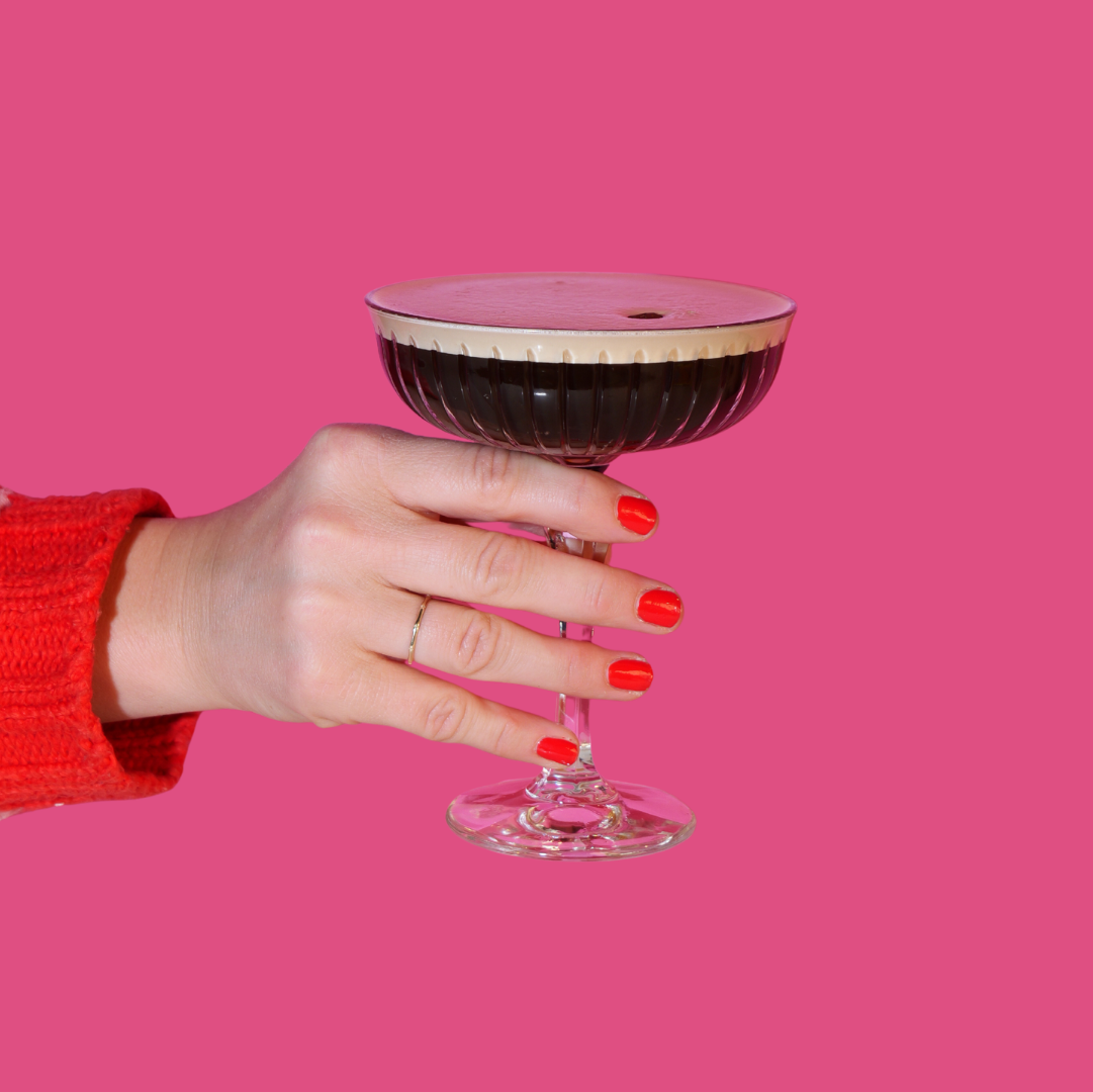 A hand with red painted nails holding an espresso martini cocktail against a pink background.