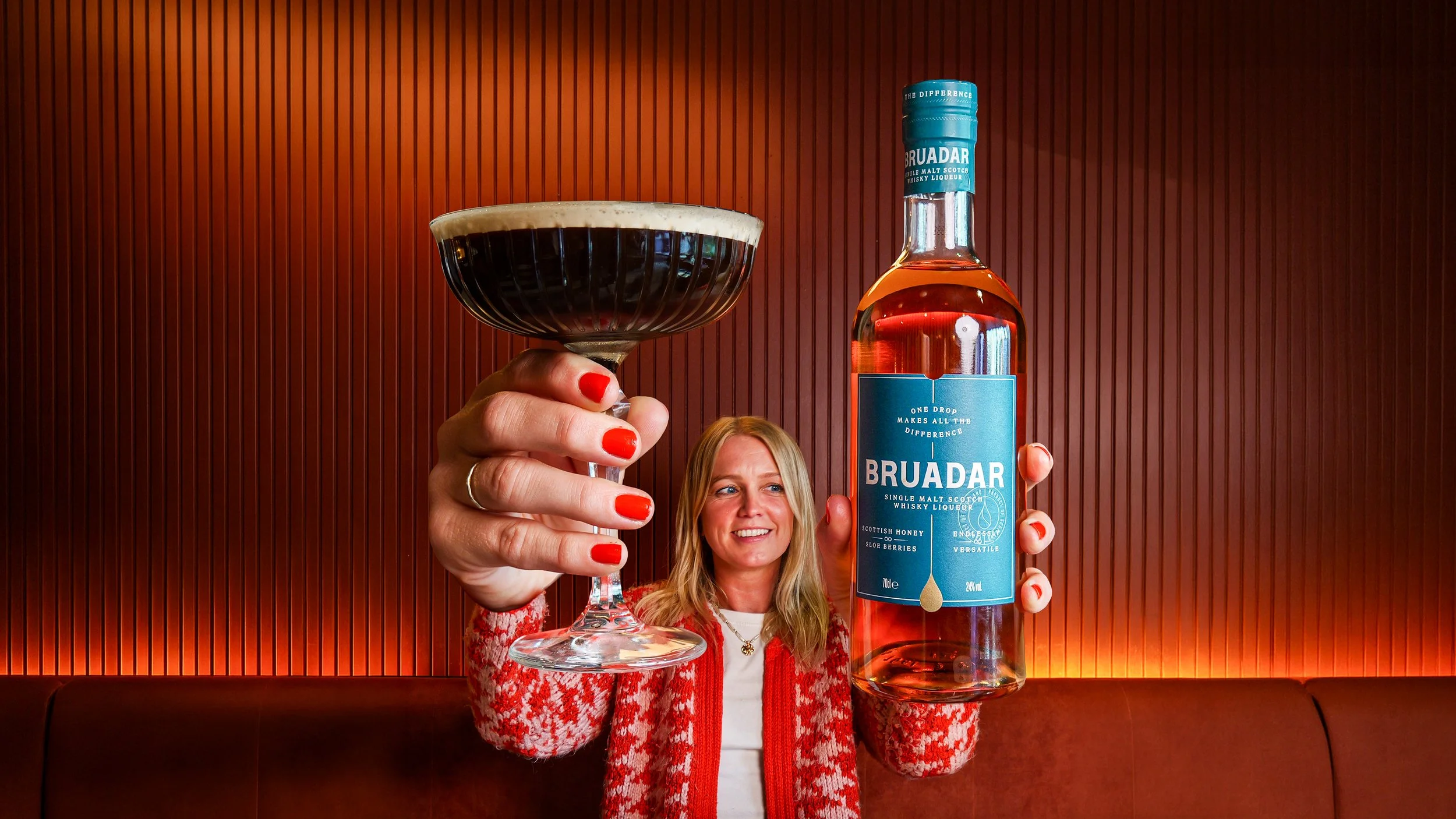 A woman with blonde hair wearing a red and white sweater holding an espresso martini and a bottle of Bruadar whisky in front of a wooden wall.
