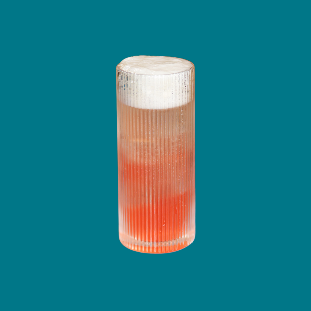 A tall, ribbed glass filled with a layered pink and orange beverage topped with frothy white foam, set against a solid blue background.