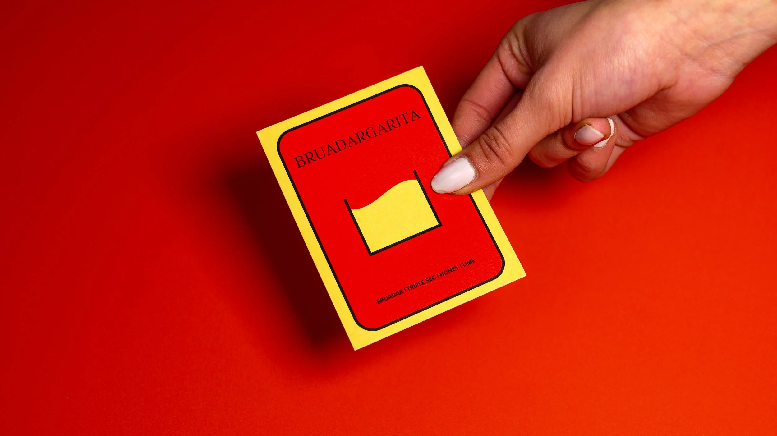 A person's hand holding a bright yellow and red drink coaster with the words 'BRUADAR' and 'BRUADARGARITA' printed on it, against a red background.