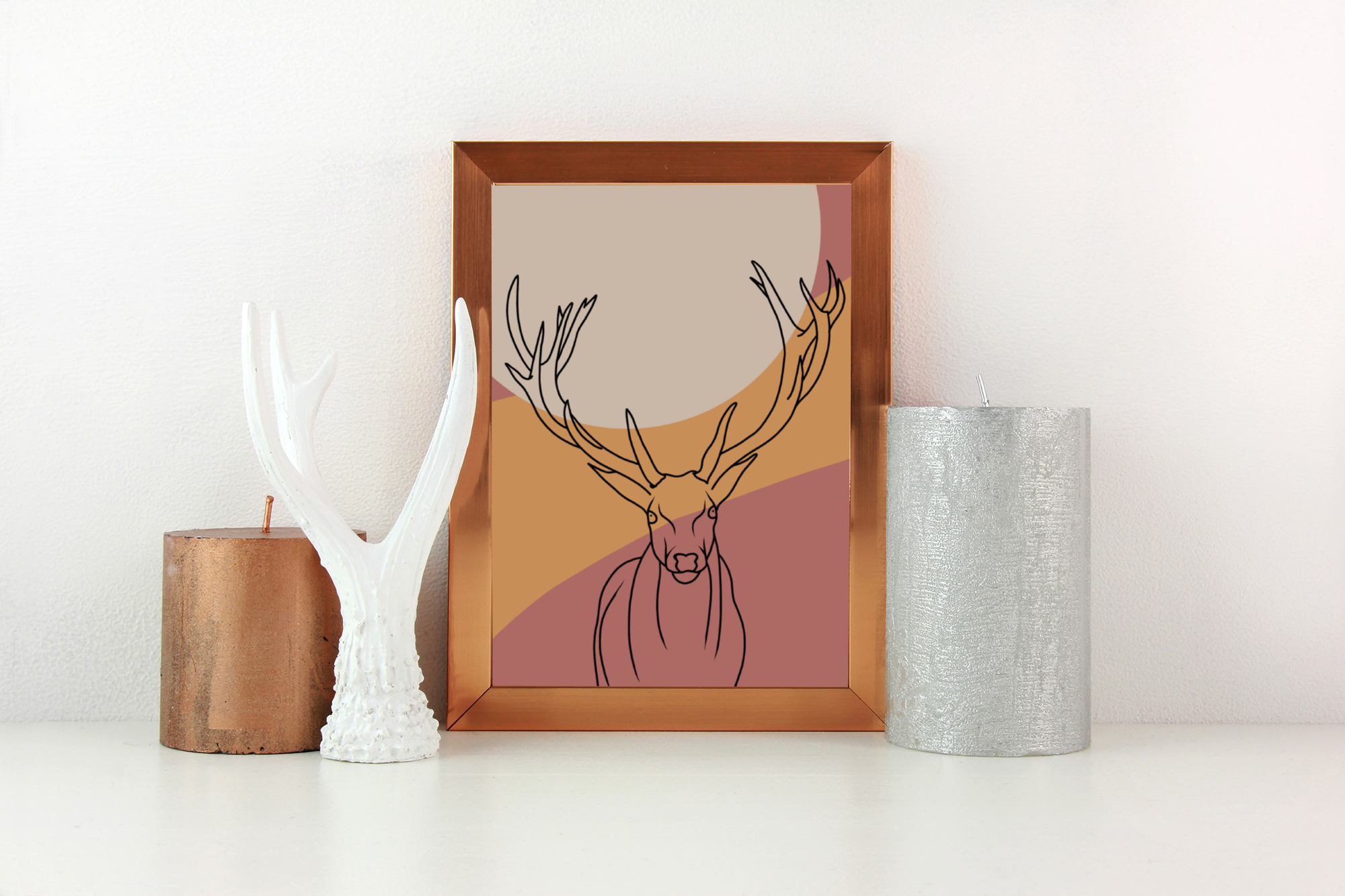 Decorative setup featuring a framed line art print of a deer, a white antler sculpture, and two textured candle holders in copper and silver on a white surface against a light-colored wall.