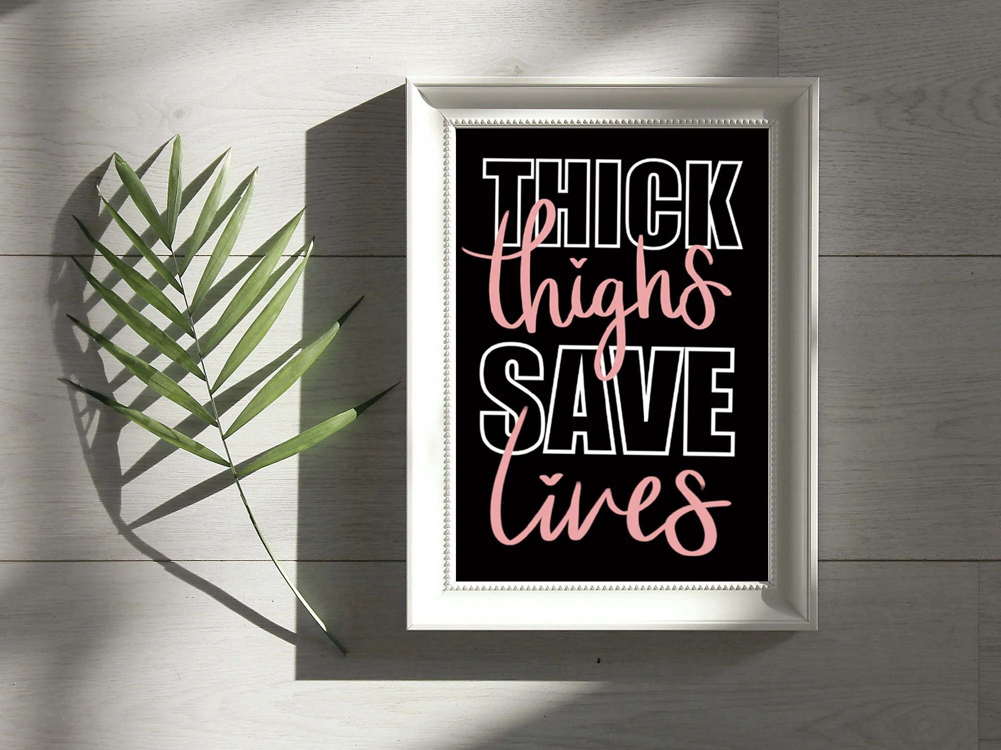 A decorative framed sign with the words "THICK thighs save lives" in pink and white text on a black background, with a green palm leaf casting a shadow on a light-colored wooden surface.