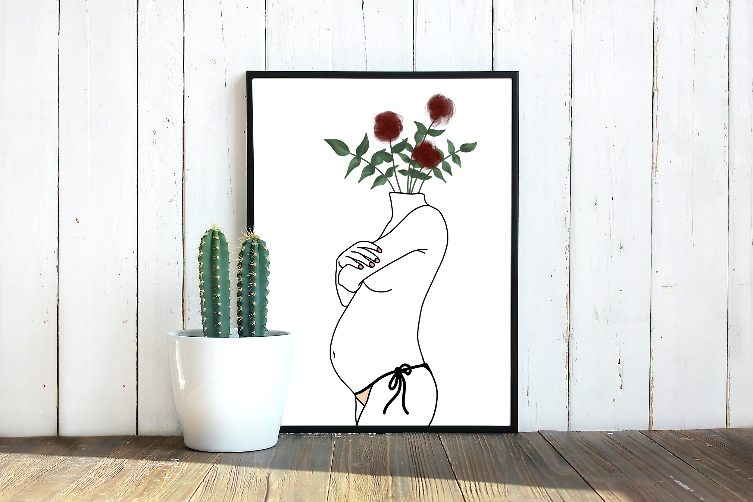 A potted cactus plant, a framed line drawing of a hand holding a flower with a red-colored flower, against a white wooden wall background.