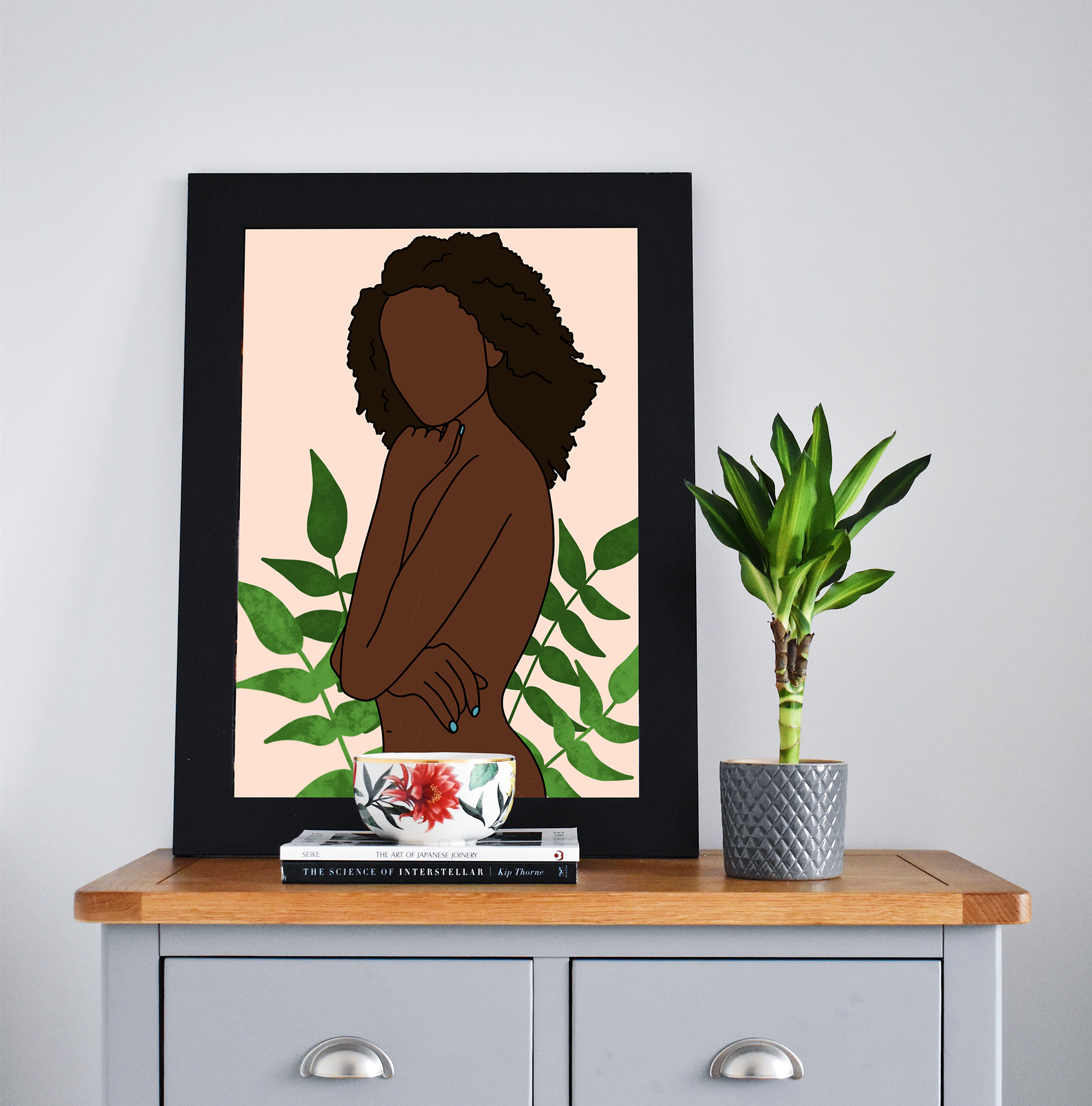A framed illustration of a faceless woman with brown skin and curly hair, surrounded by green leaves, resting on a table near a stack of books, a potted plant, and a bowl with a floral design.