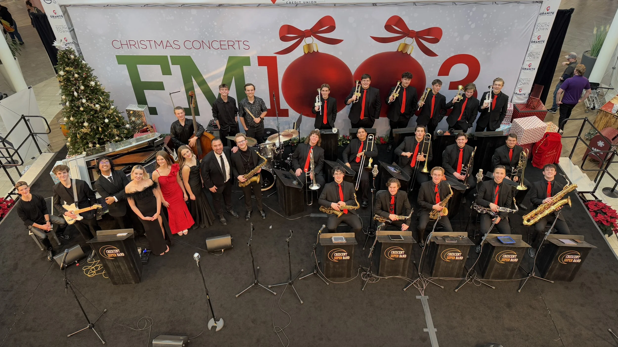The Crescent Super Band on stage in black suits and red ties, horns raised, in front of a large FM100.3 Christmas Concerts backdrop with a decorated tree beside them.