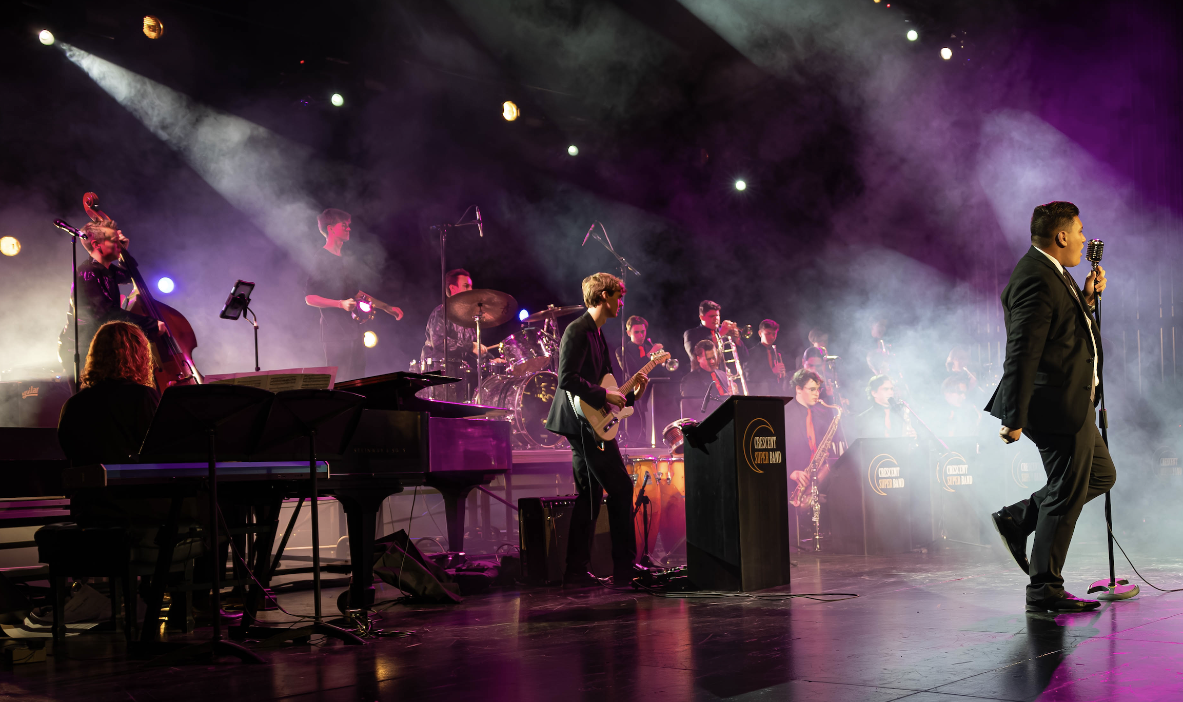 Crescent Super Band performing live onstage with a vocalist, guitarist, rhythm section, and horn section under colorful concert lights.