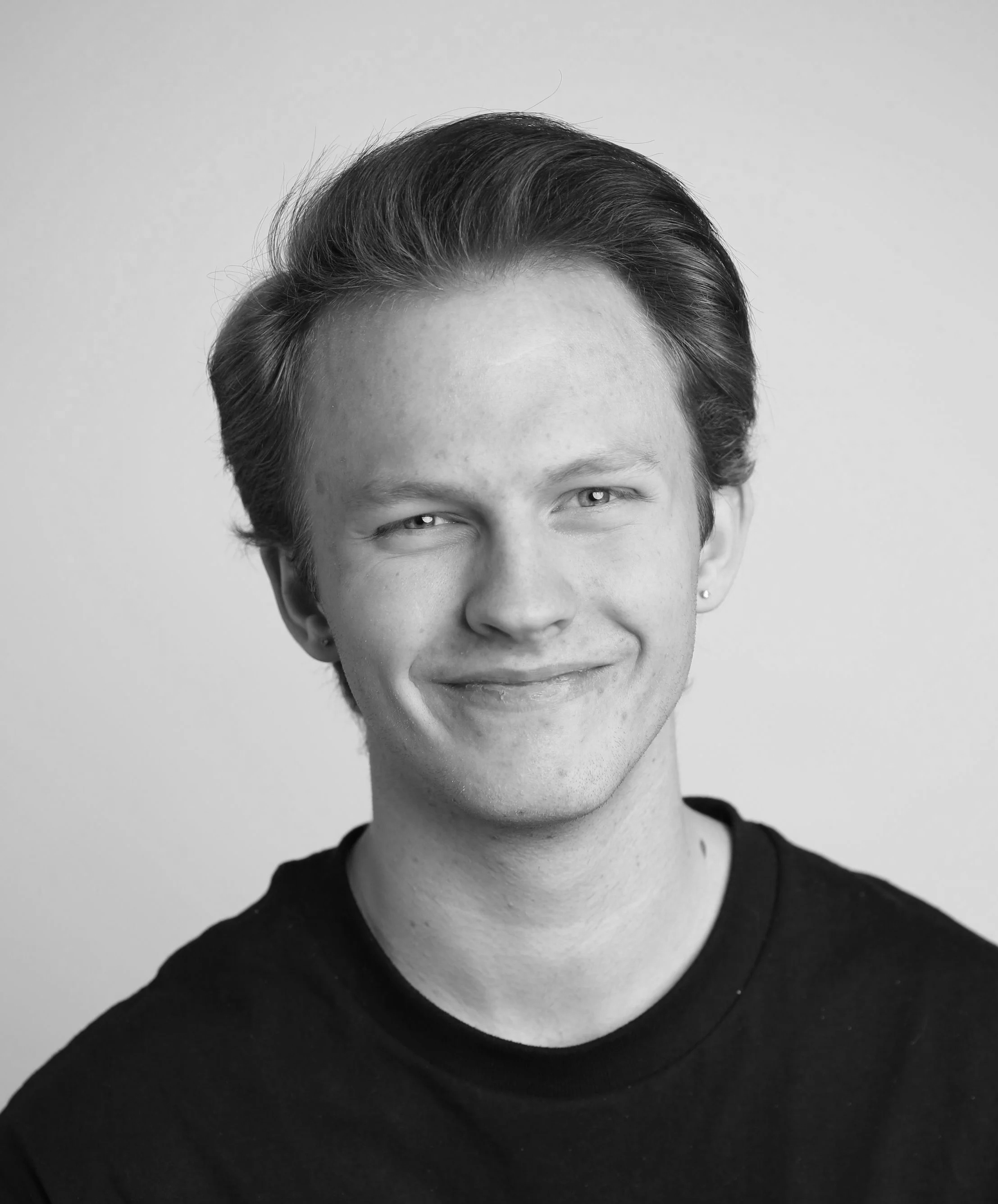 Black and white portrait of a young man with light skin, wearing round glasses, short wavy hair, and a black shirt, smiling slightly against a plain background.