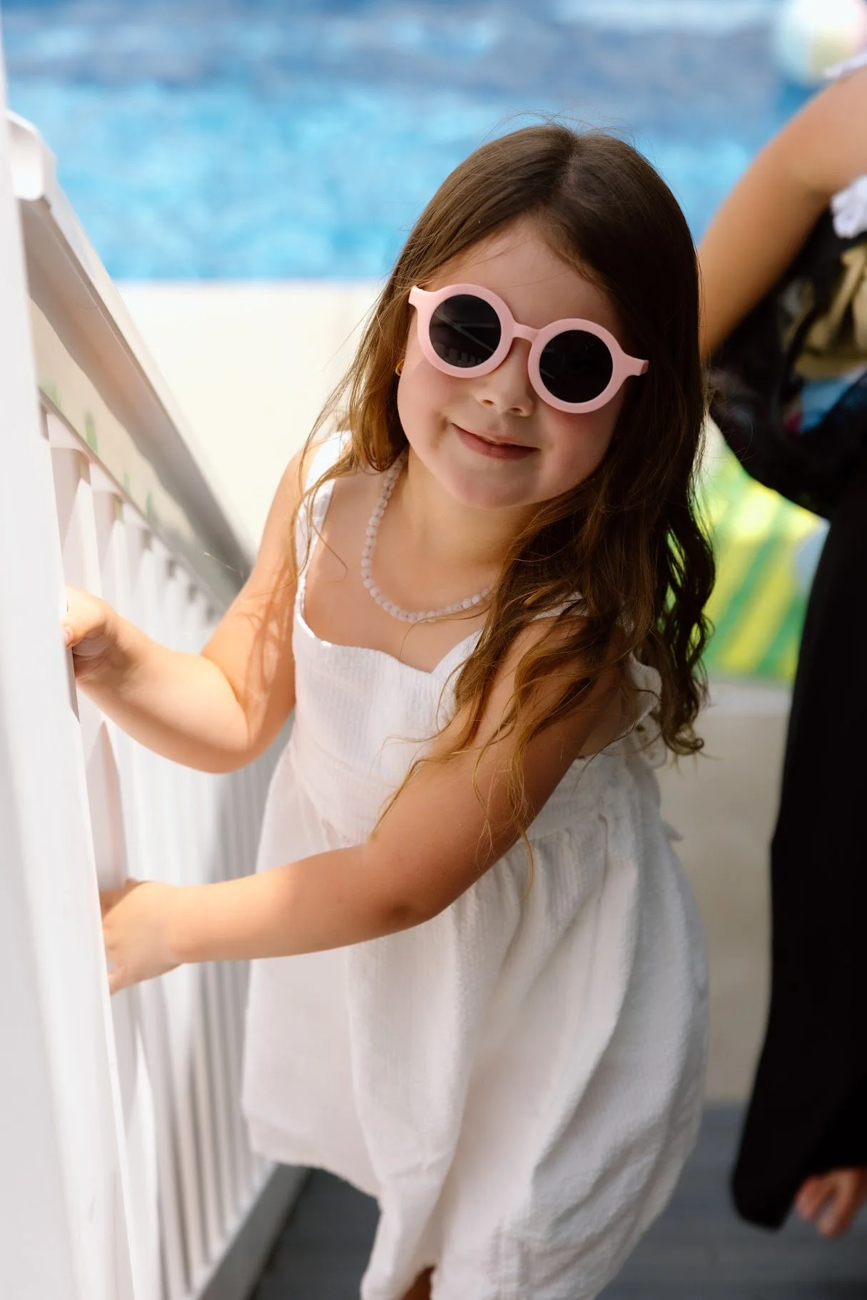 Young girl in a white dress with a pearl necklace, wearing pink sunglasses, standing by a white fence near a swimming pool.
