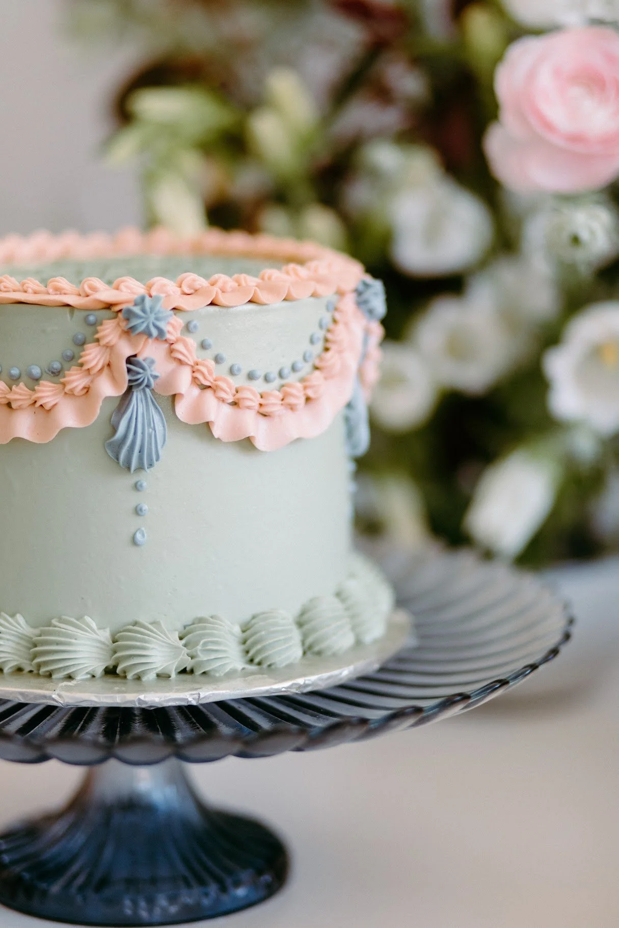 A decorated cake with pastel pink and blue icing, floral decorations, and topped with a pink and blue border, placed on a black cake stand with a white base.