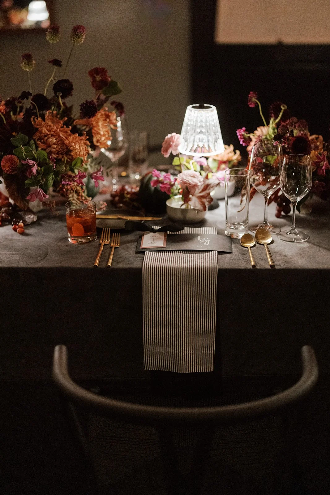 A formal dining table decorated with pink and red flowers, a small table lamp, and elegant glassware, set for a meal.