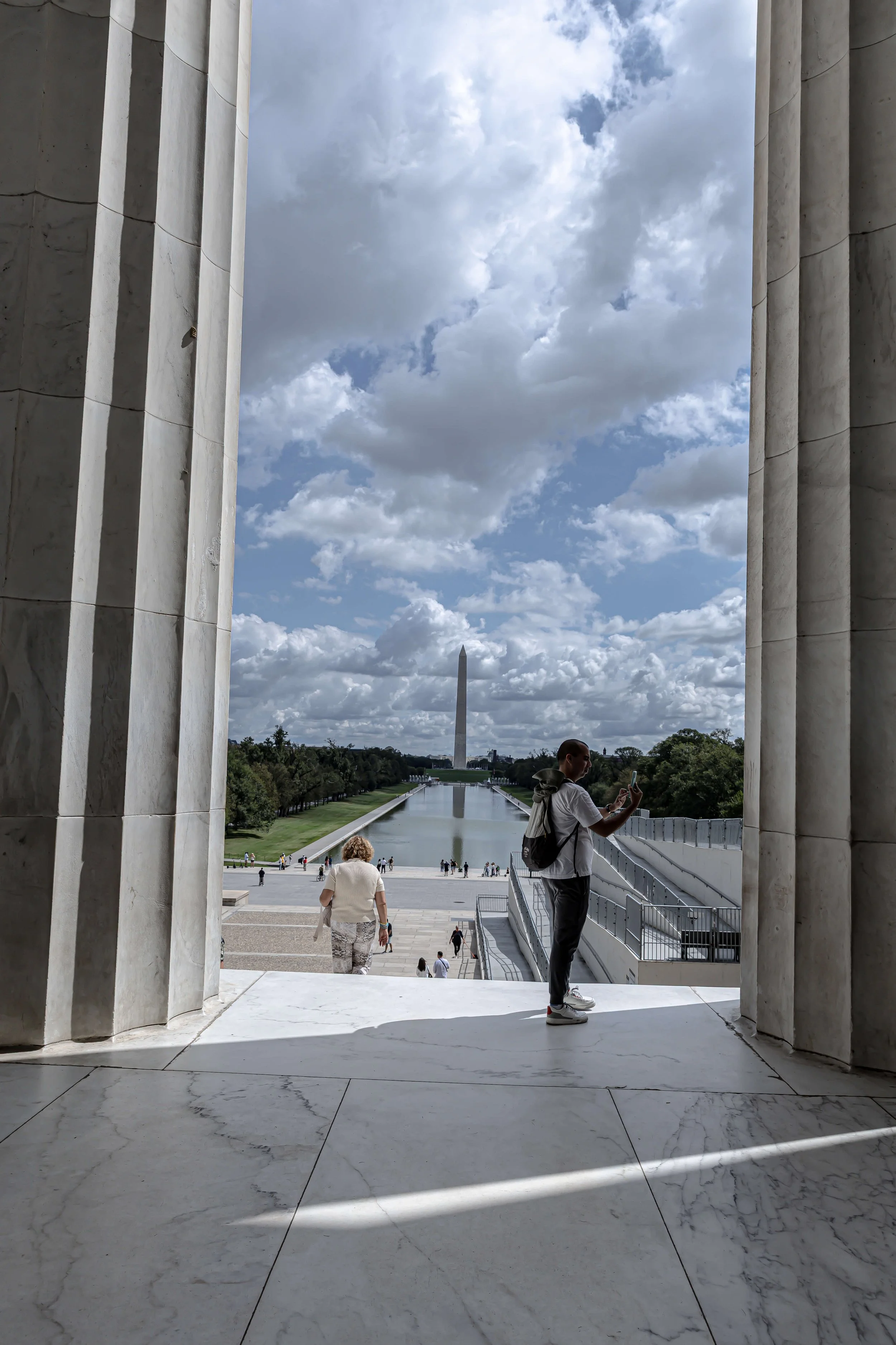 View through the Lincoln Memorial in Washington, D.C., featuring the Reflecting Pool and the Washington Monument in the distance under a partly cloudy sky, with visitors walking and taking photos.