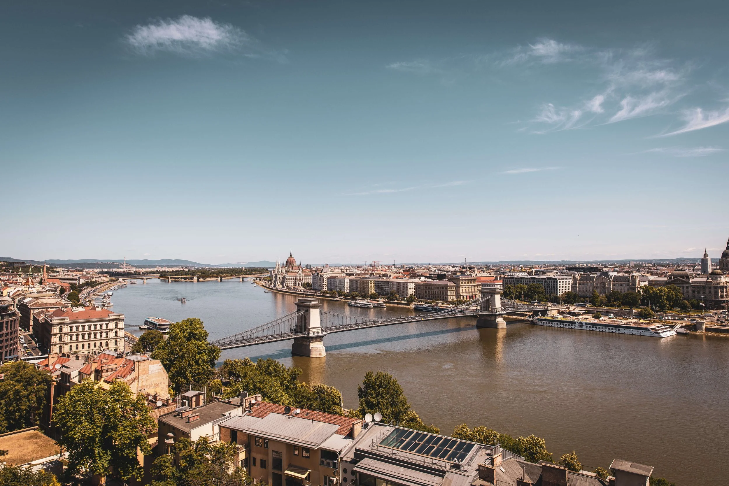 A wide cityscape of Budapest, Hungary, with the Danube River running through the center. The Chain Bridge crosses the river in the foreground, and the Hungarian Parliament Building and other historic structures are visible along the riverbank. The sk