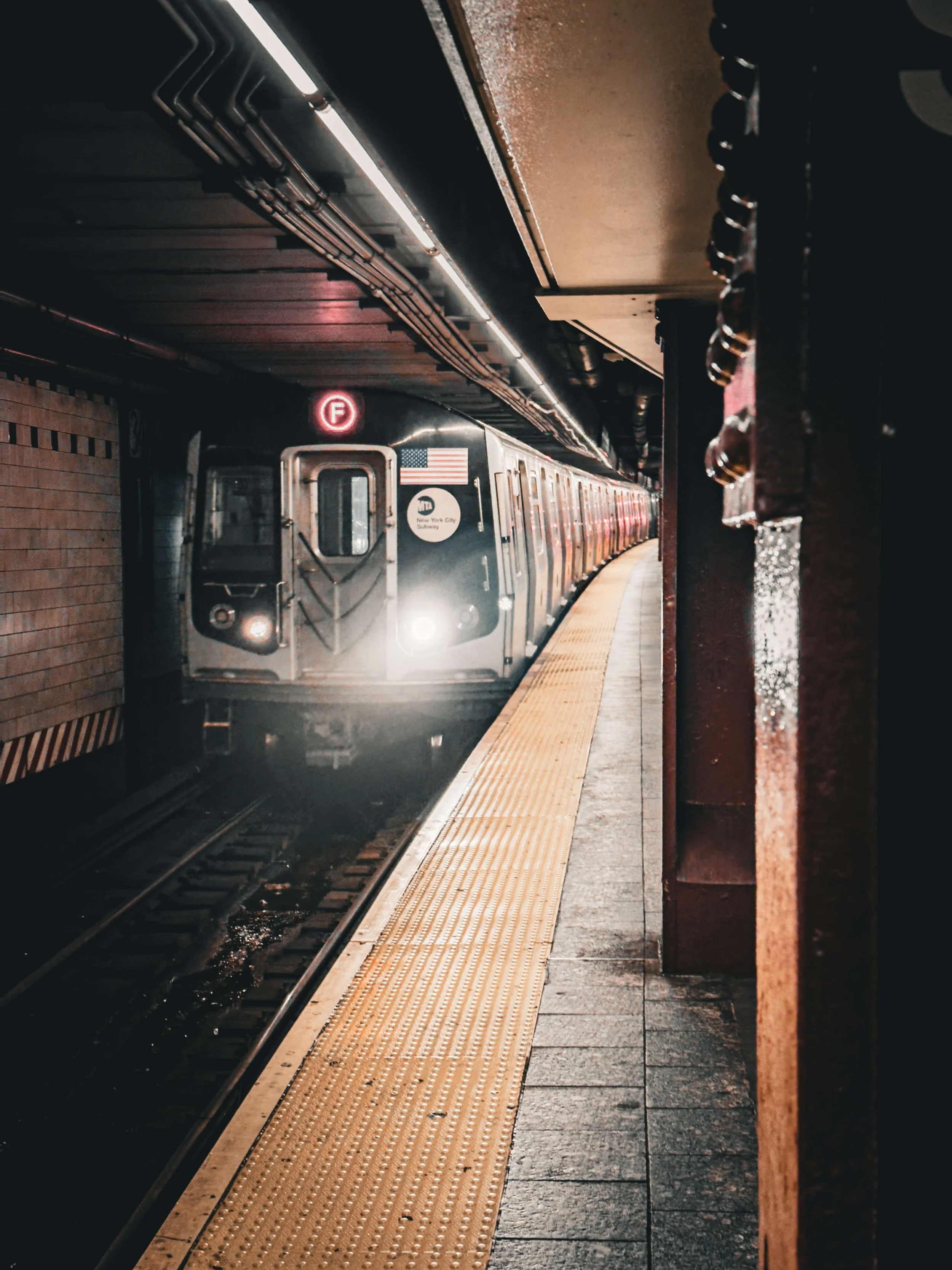 An NYC subway train approaching a station platform at night, with a red 'F' subway line sign illuminated above the train.