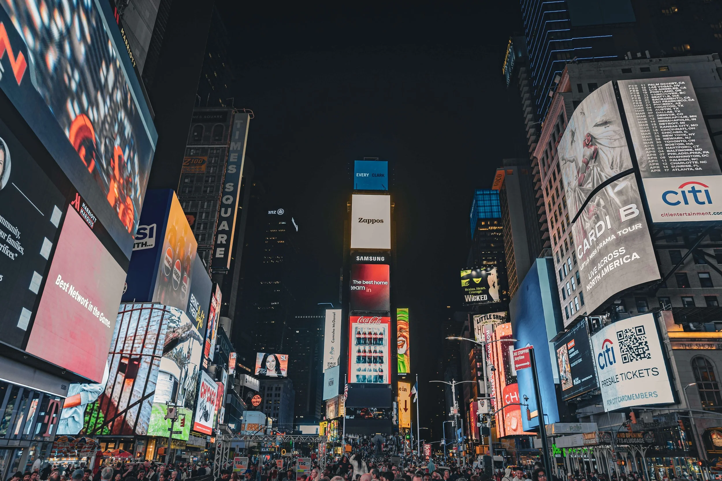 Nighttime scene of Times Square, New York City, with bright digital billboards and advertisements, and people walking below.